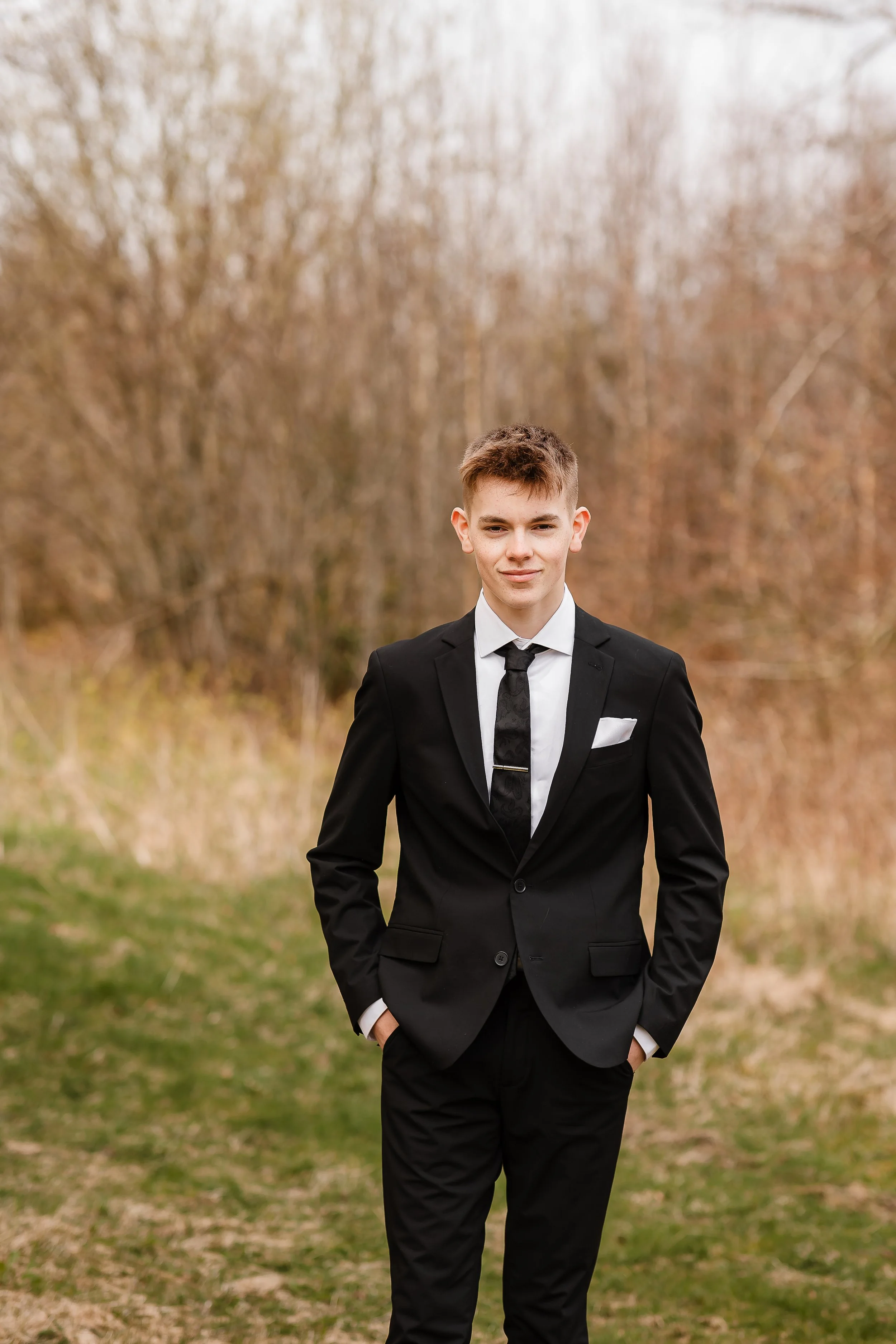 Ung mann i svart dress, hvit skjorte og svart slips står ute i naturen, med trær som har høstfarger i bakgrunnen.