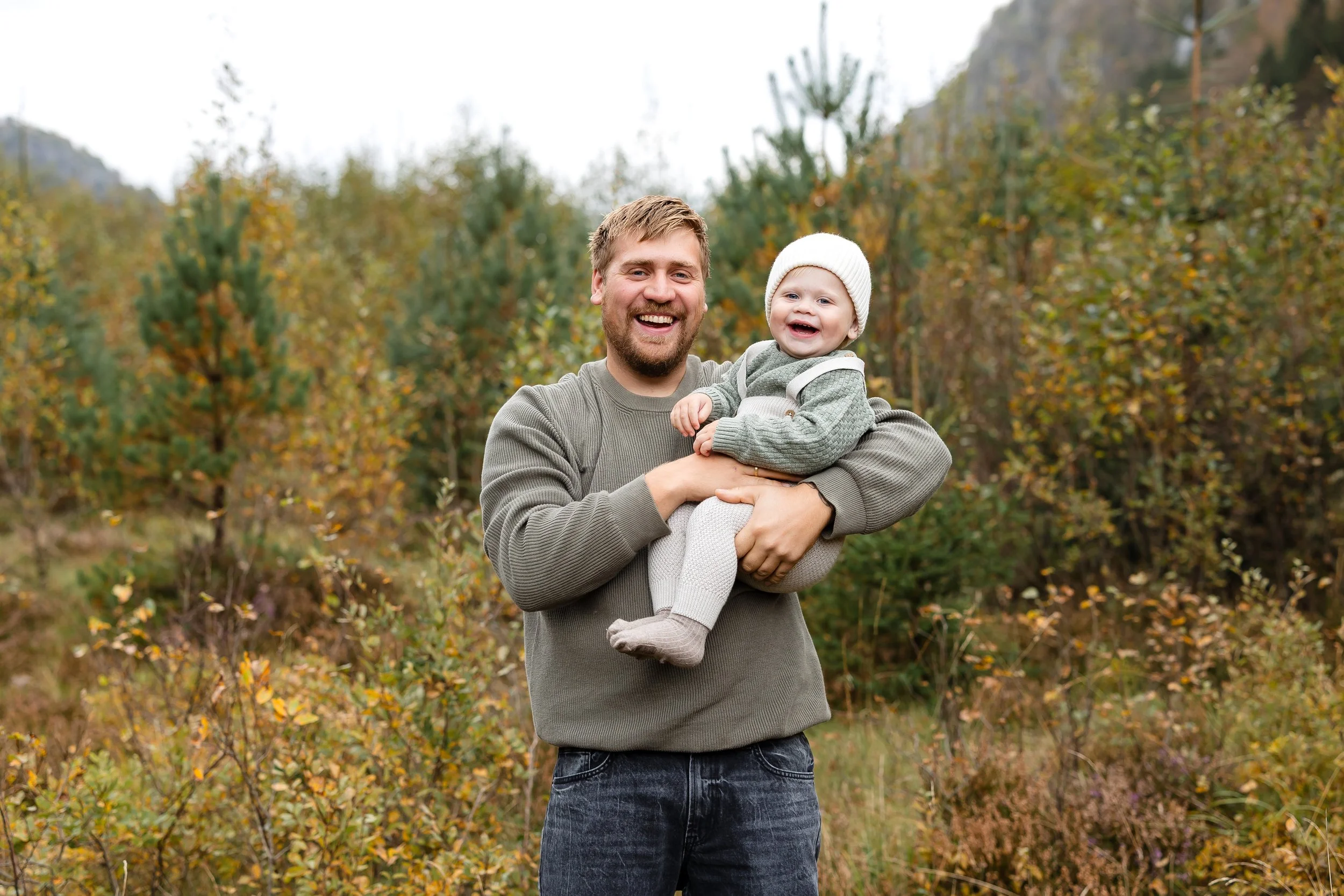 En voksen mann med skjegg smiler og holder en liten jente med hvit lue og grågenser i armene. De står i en skog med høstfargede trær.