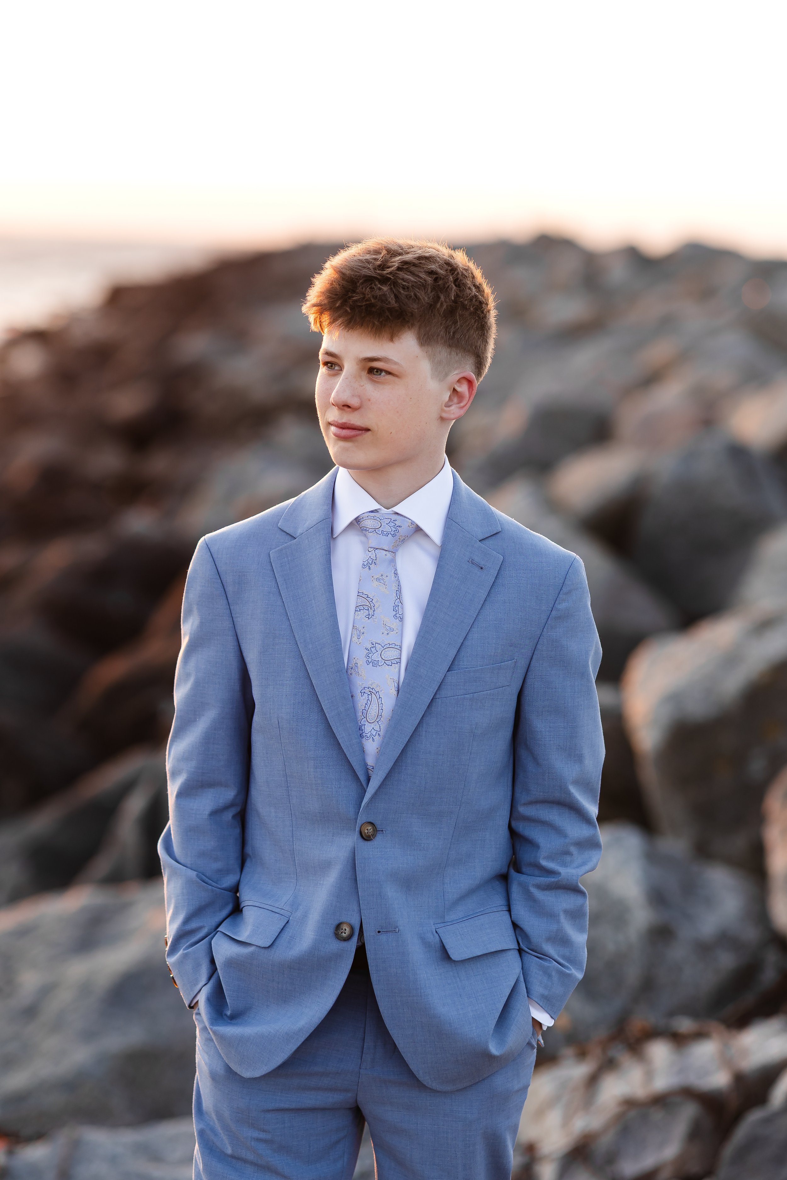 Ung mann i blå dress står på en steinete strand ved solnedgang.