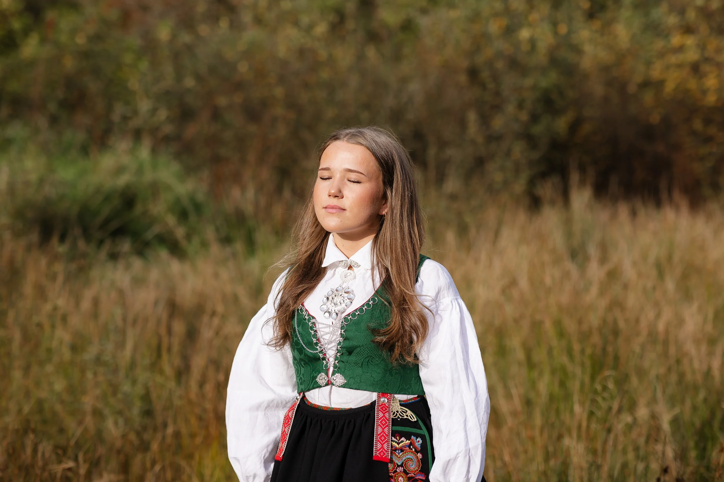 Ung kvinne i tradisjonell norsk bunad står i en åker med høstfarger.