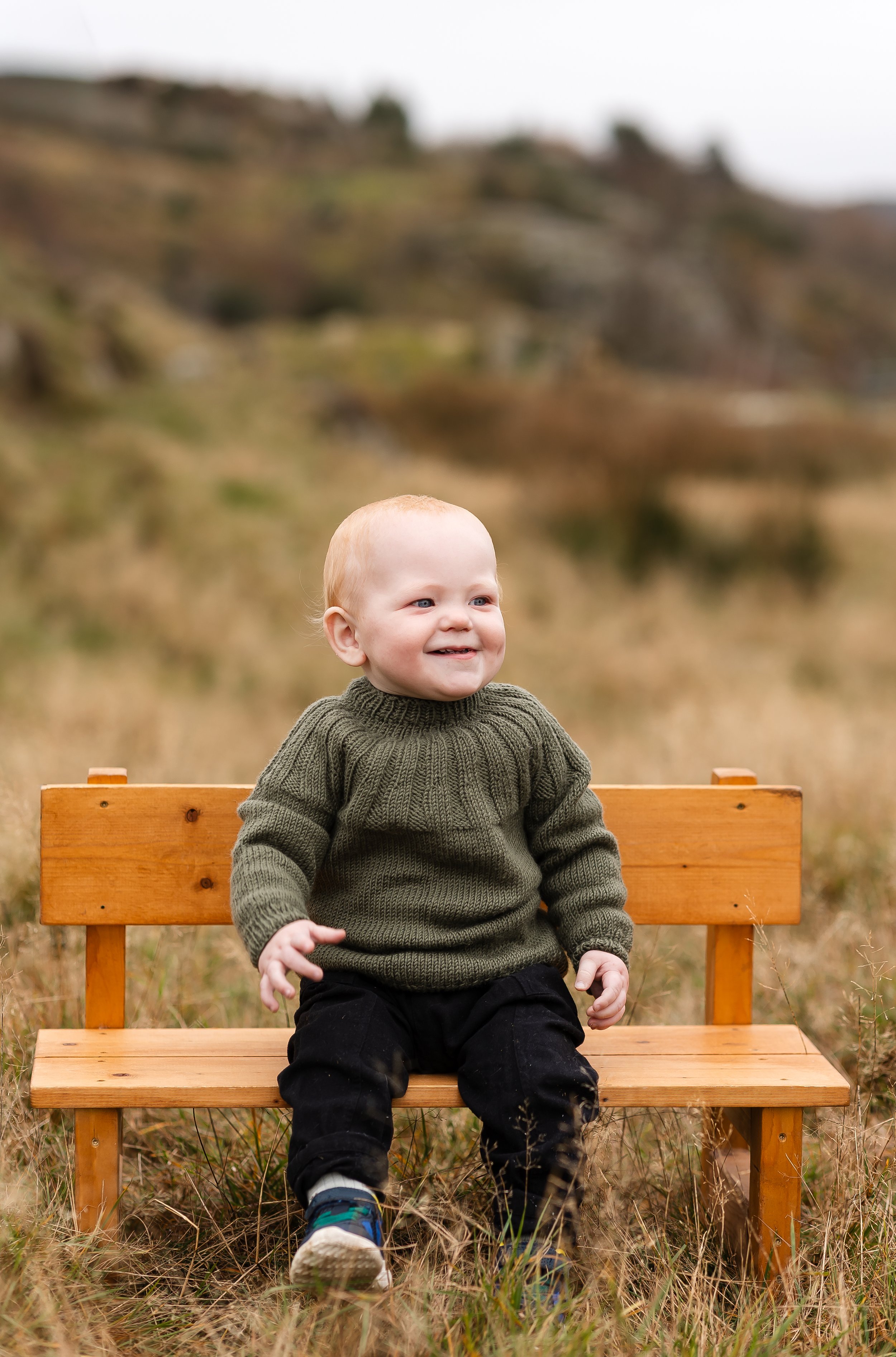 En smilende liten gutt med lyst hår som sitter på en liten trebenk i en utendørs setting med gress og bakgrunn av naturlandskap.