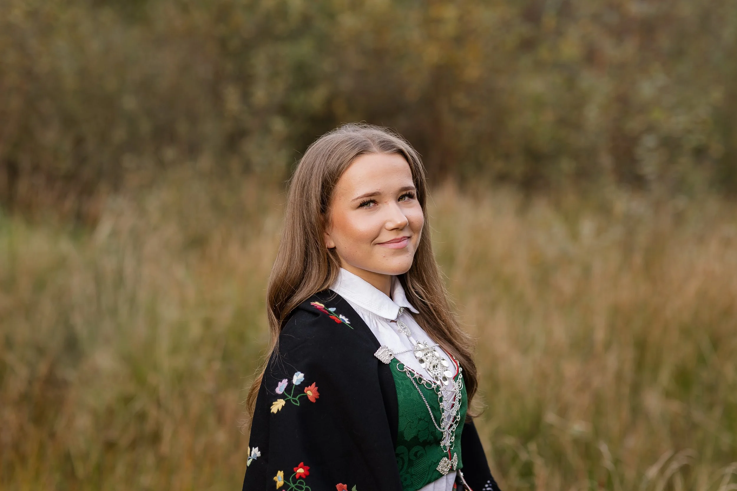 Ung kvinne i tradisjonell norsk bunad står i en åpen skog eller åker med høstfarger i bakgrunnen, smilende og ser mot kamera.