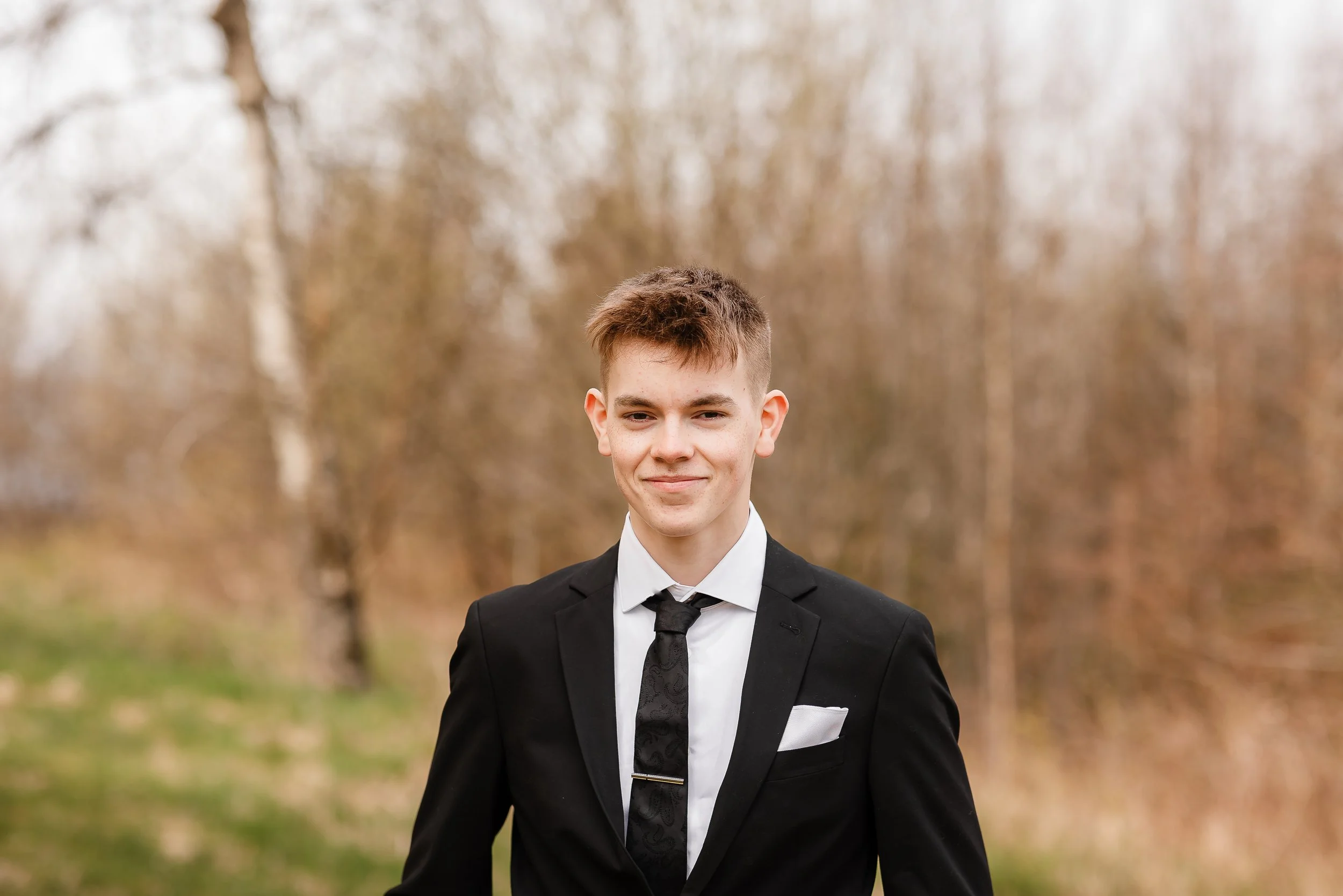 Ung mann i svart dress, hvit skjorte og svart slips som står utendørs på en åker, med bartrær i bakgrunnen, smilende mot kamera.