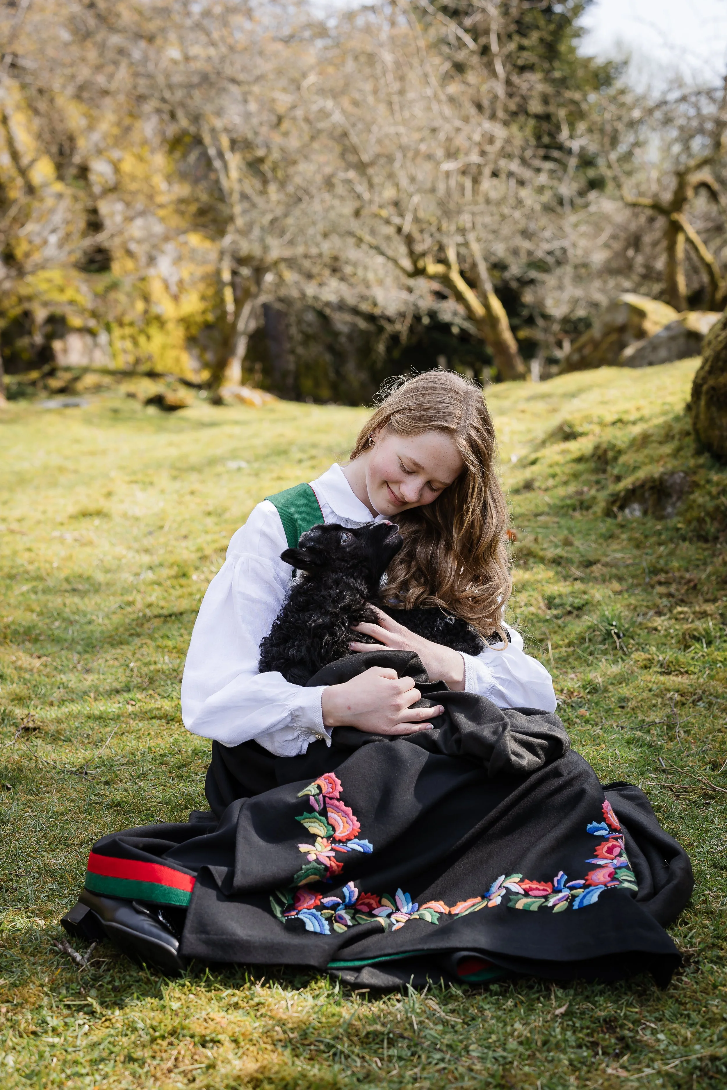 En jente med langt, krøllete hår sitter på gresset i en eng, holder en liten svart valp og koser med den i et naturskjønt landskap.