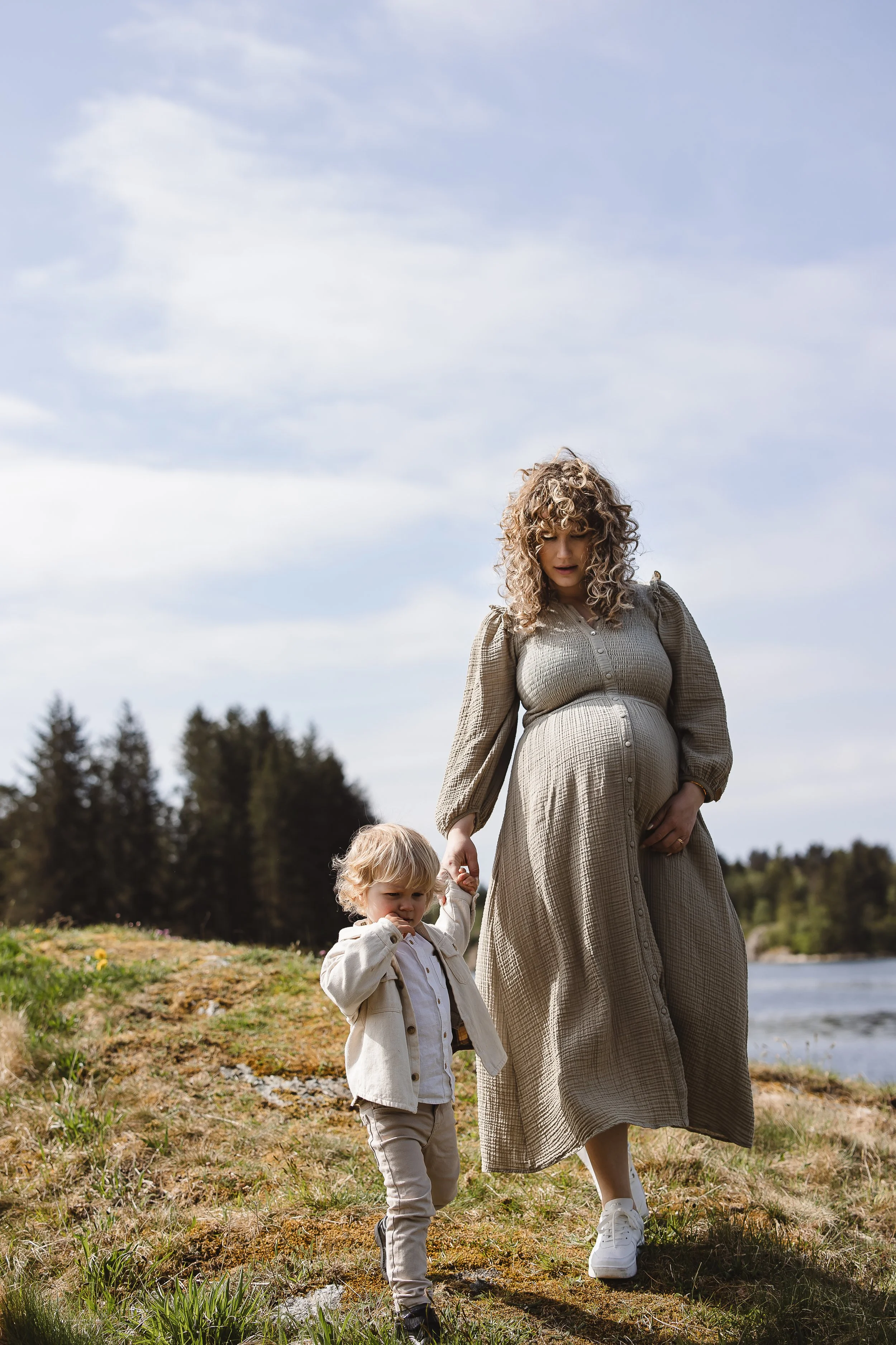 En gravid kvinne går hånd i hånd med et lite barn ved en elv under en gråværsdag.