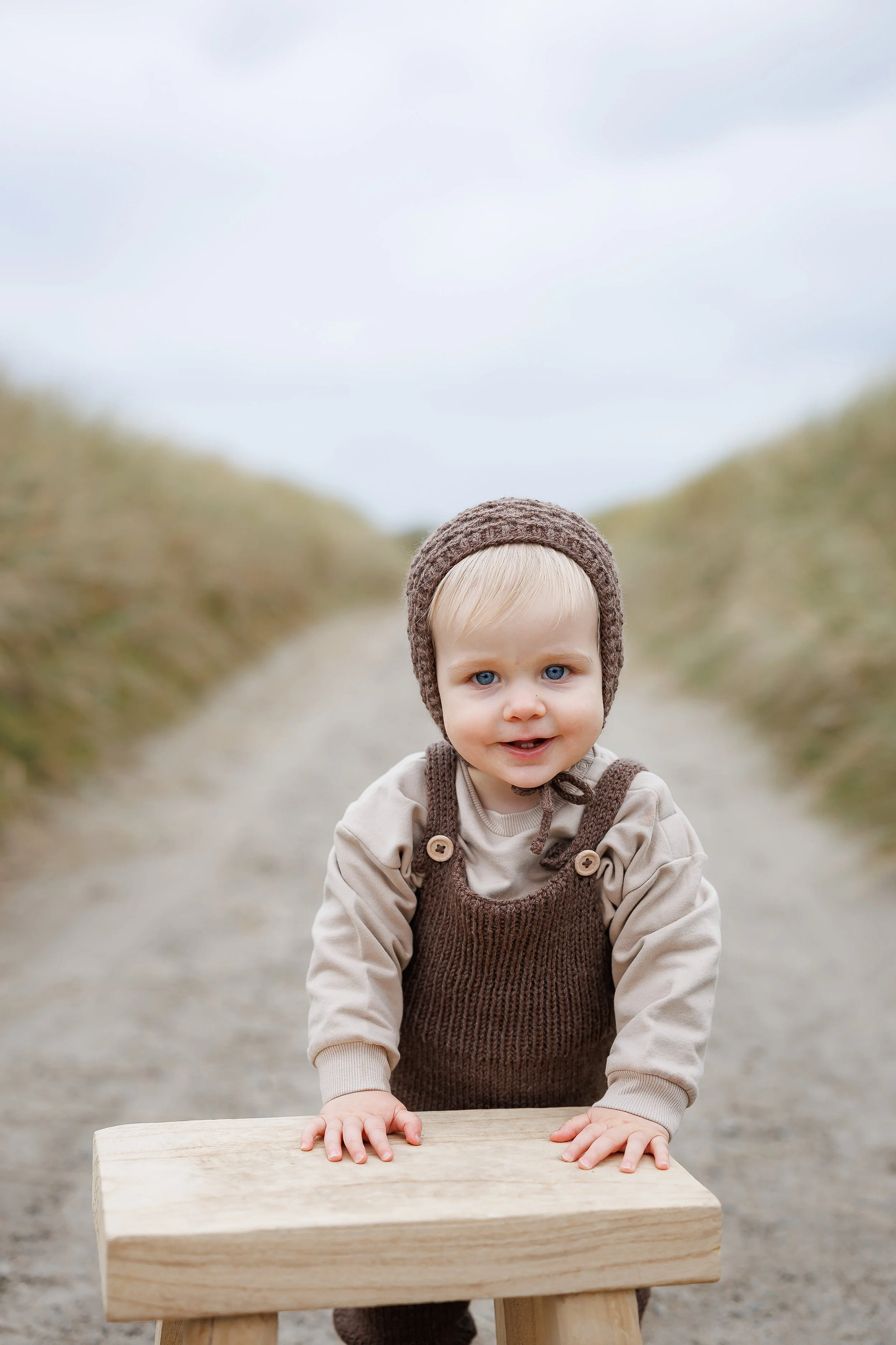 Liten gutt som står på en trebenk på en grusvei, iført beigegenser, brune luer og spansker, med blå øyne og lyst hår, ute i naturen.