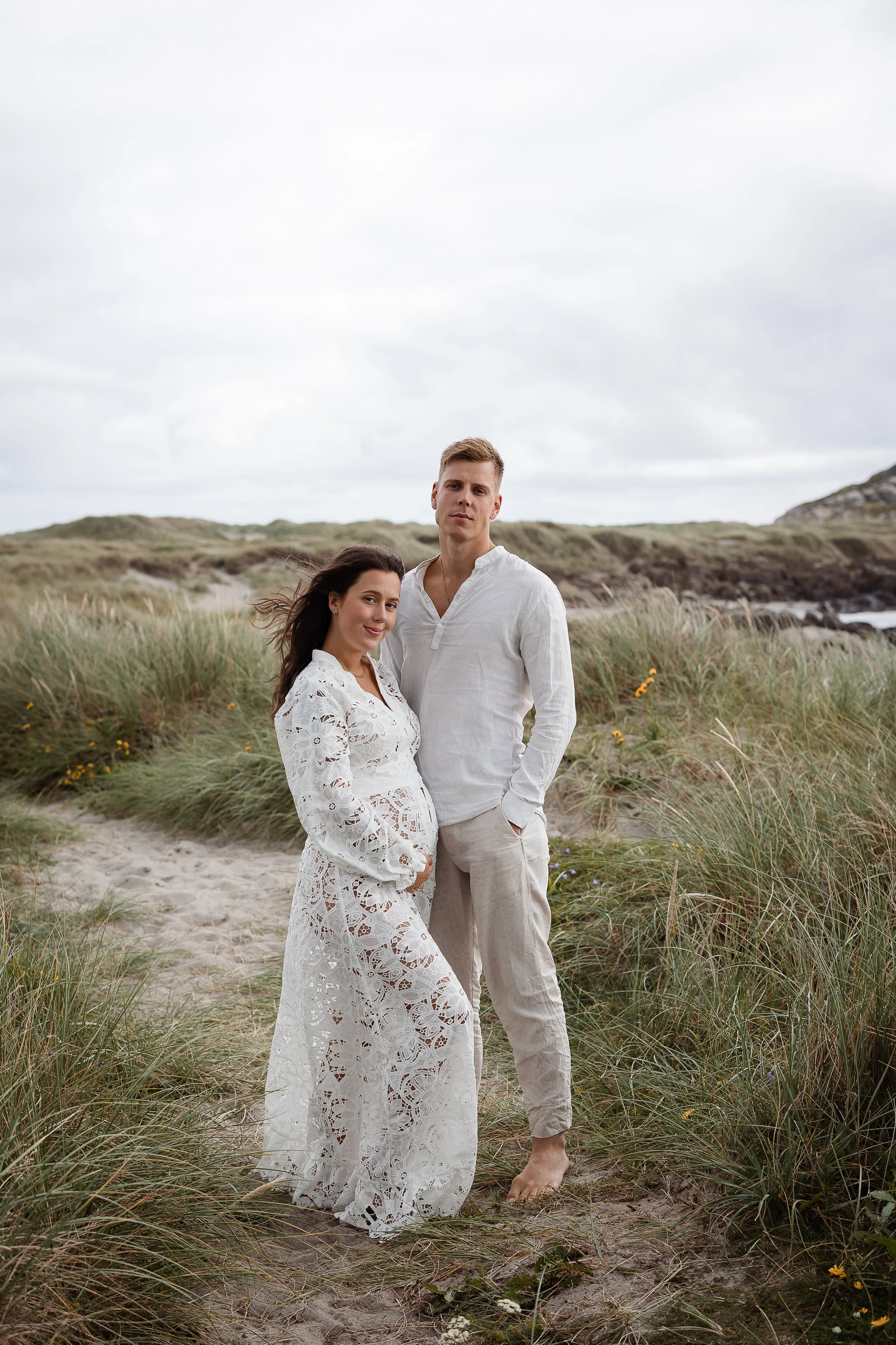 Et par i hvite klær står på en sandstrand med gress og fjell i bakgrunnen.