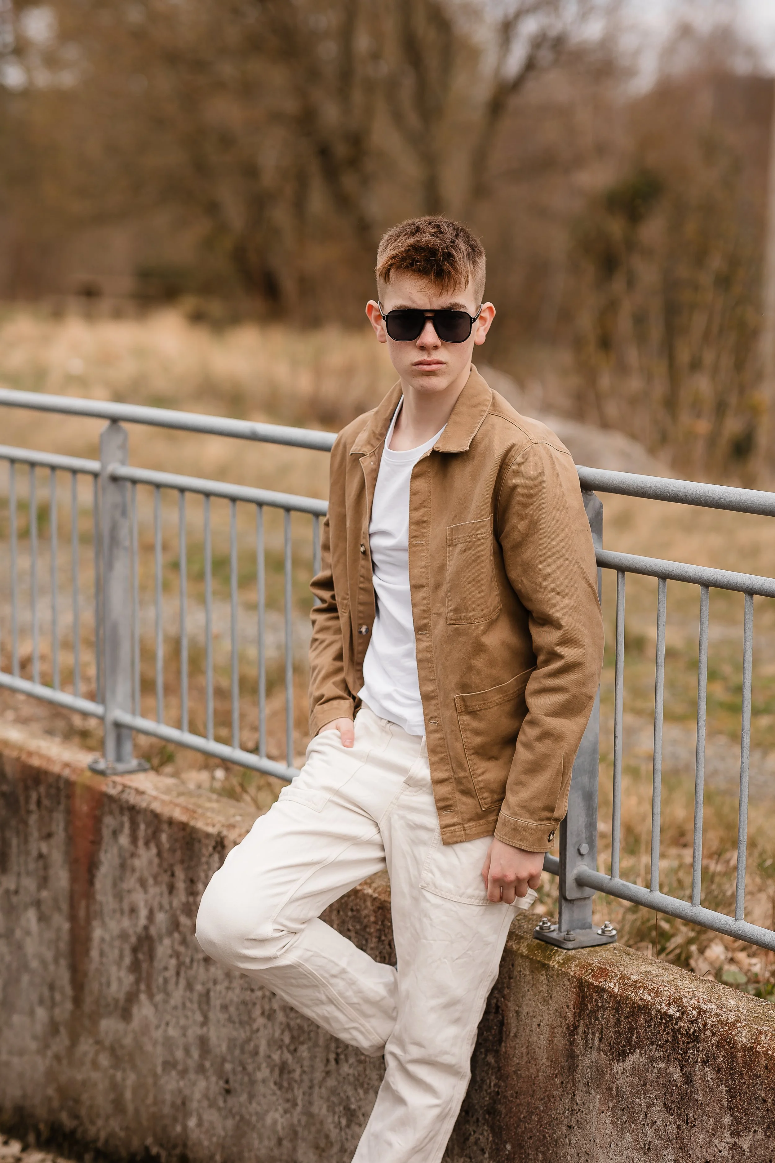 Ung mann med solbriller i beige jakke og hvite bukser står ved en metal railing i en park med høstfarger i bakgrunnen.