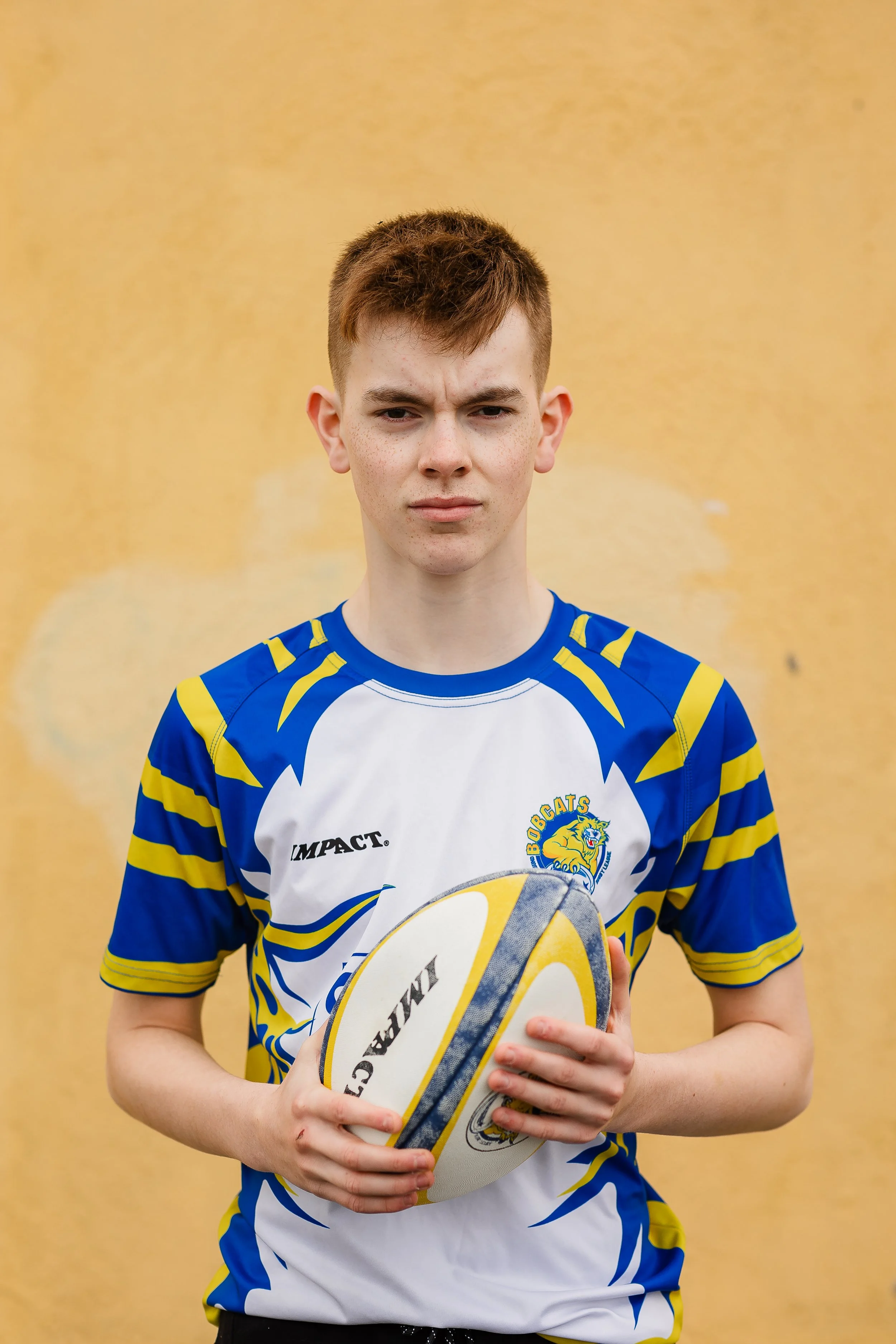 Ung gutt i rugbydrakt holder en rugbyball, står foran en gul vegg.