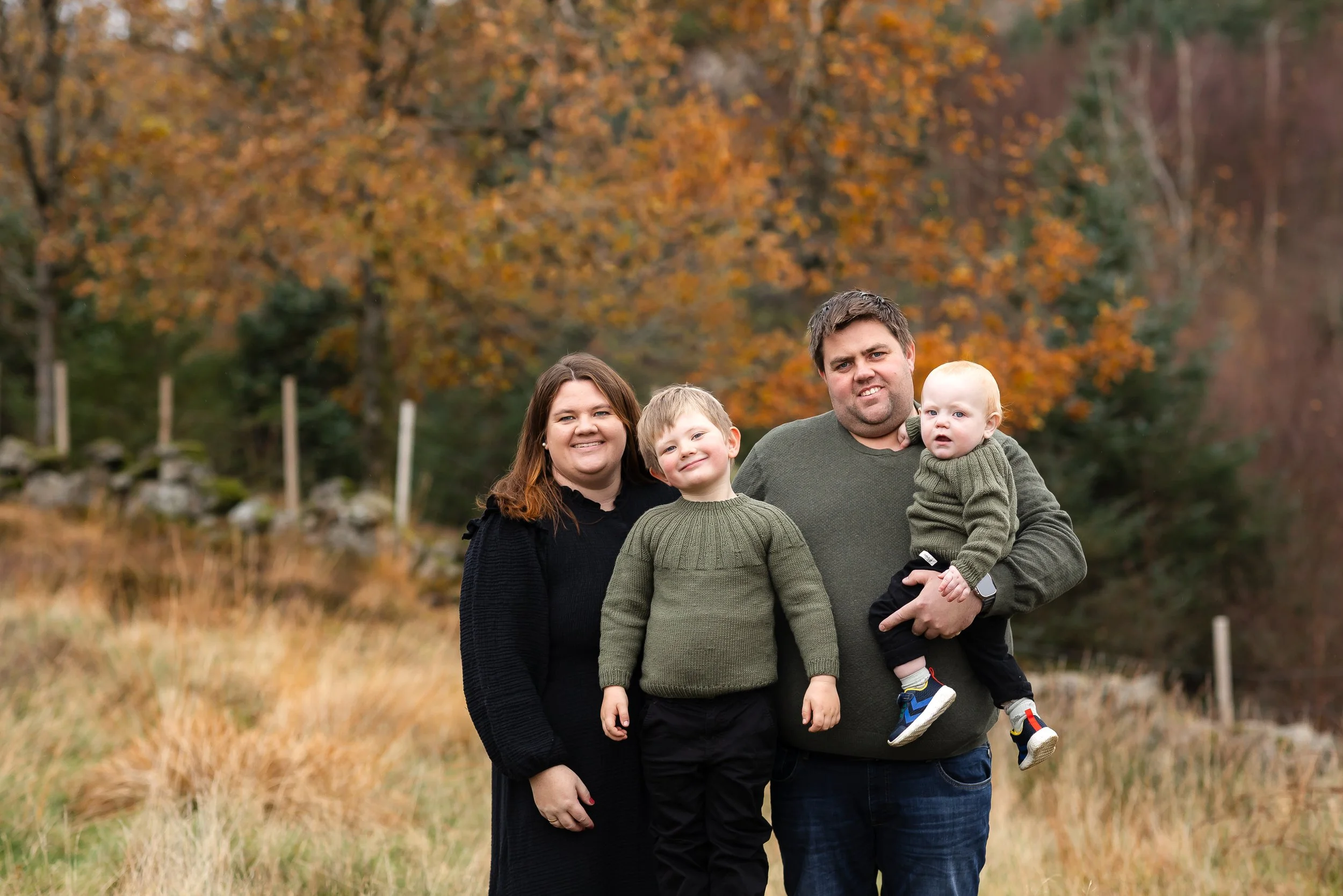 En familie på fire står ute i høstlandskap, med høstfargede trær i bakgrunnen.