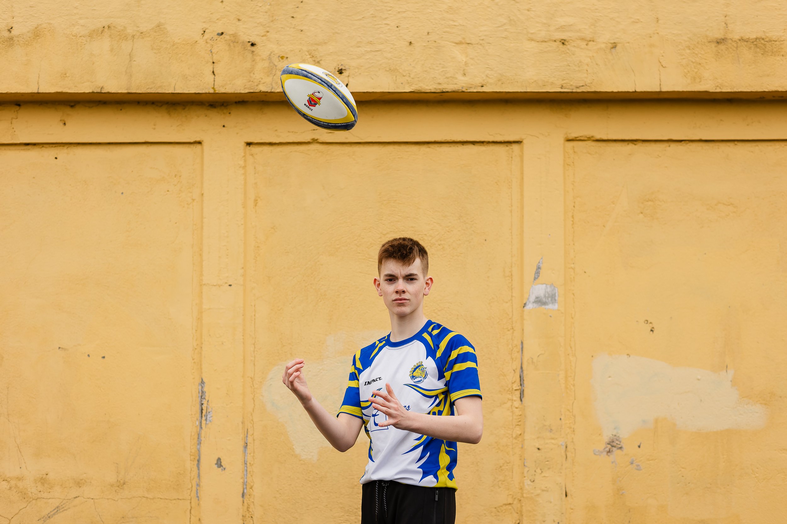 En ung mann i en blå og hvit rugby-trøye står foran en gul vegg, med en rugbyball i lufta over hodet.