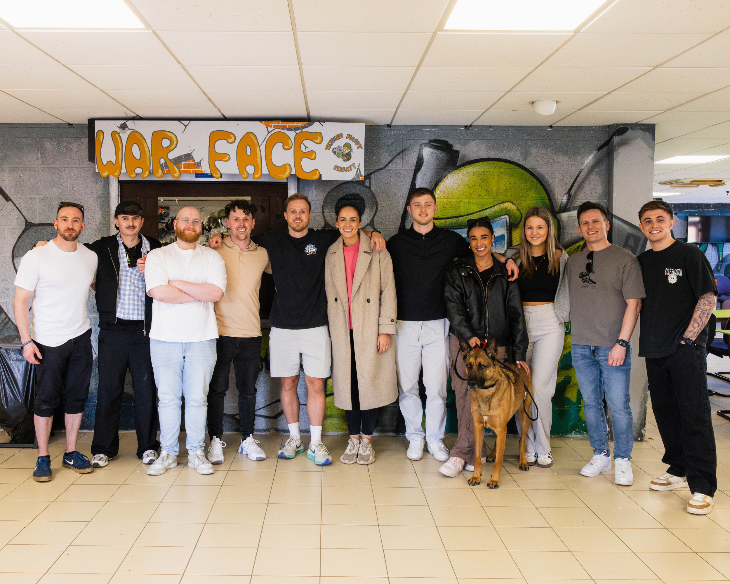 A group after their Trigger Happy event posing for a photo. They are casually dressed and smiling. Behind them is a graffiti-style mural with the words 'WAR FACE.' A dog is present, standing in front of one of the people.