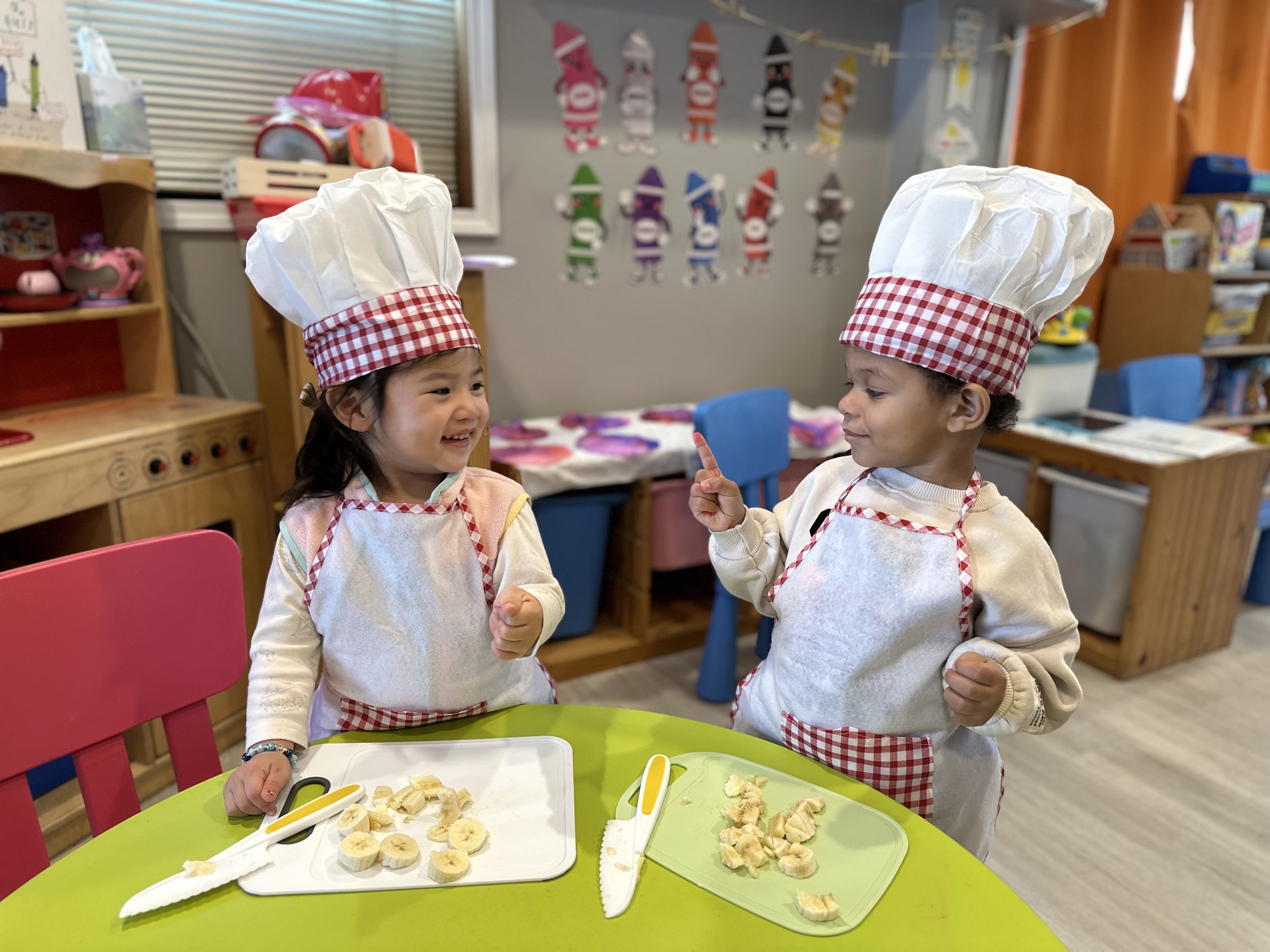 Two young children wearing chef hats and aprons, standing at a green table with sliced bananas, engaging in a playful cooking activity in a colorful classroom.