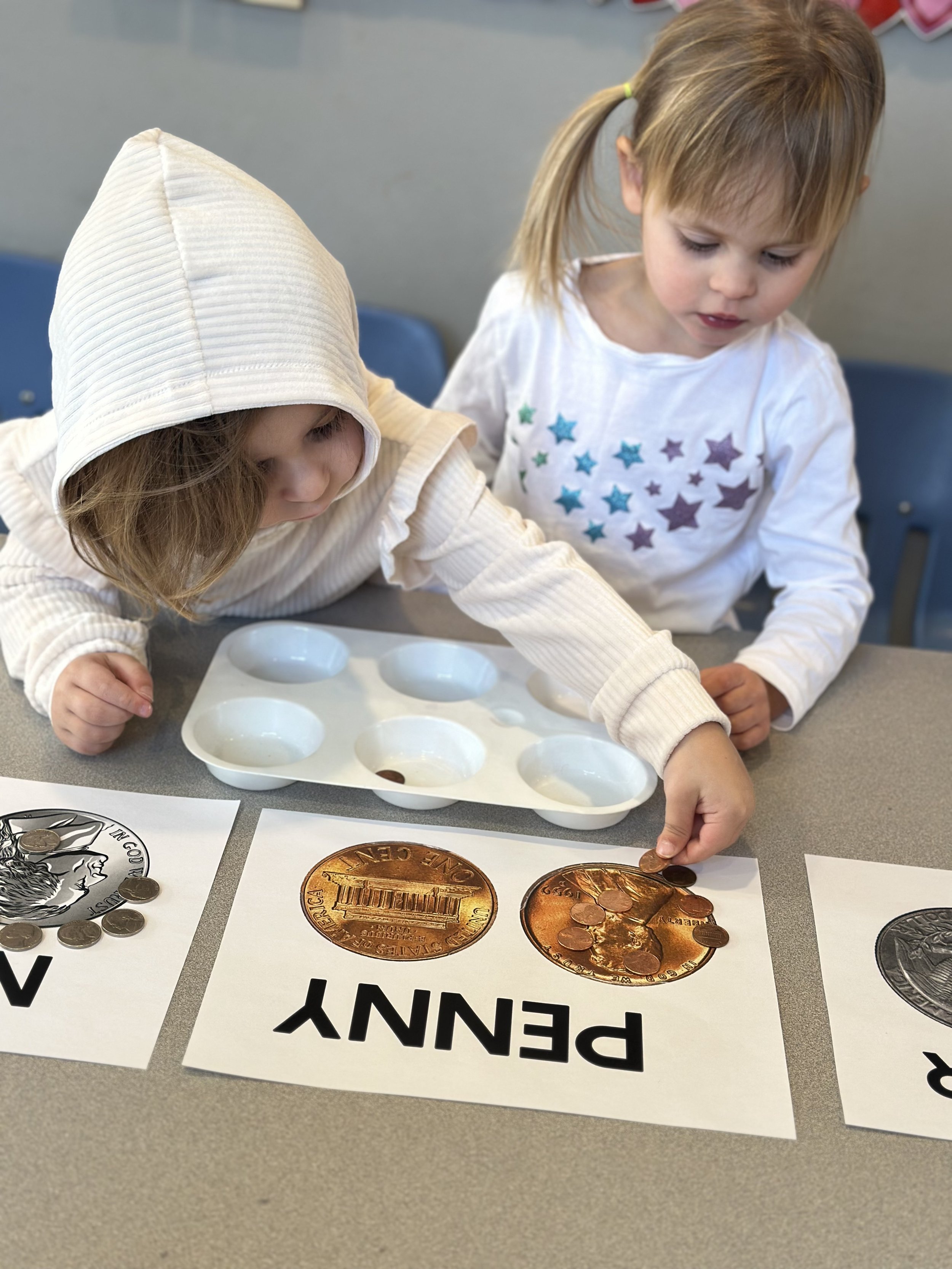 Two young children working with coins, placing coins into penny-themed educational posters on a table at a classroom activity.