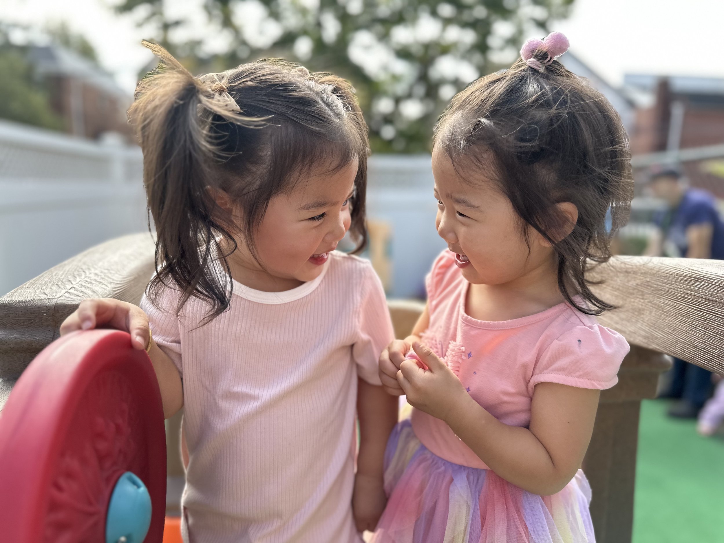 Two young girls are smiling and laughing together outdoors, holding hands near a playground. One girl is wearing a light pink top, and the other girl is wearing a pink dress with a rainbow-colored skirt. They appear to be having fun and enjoying each