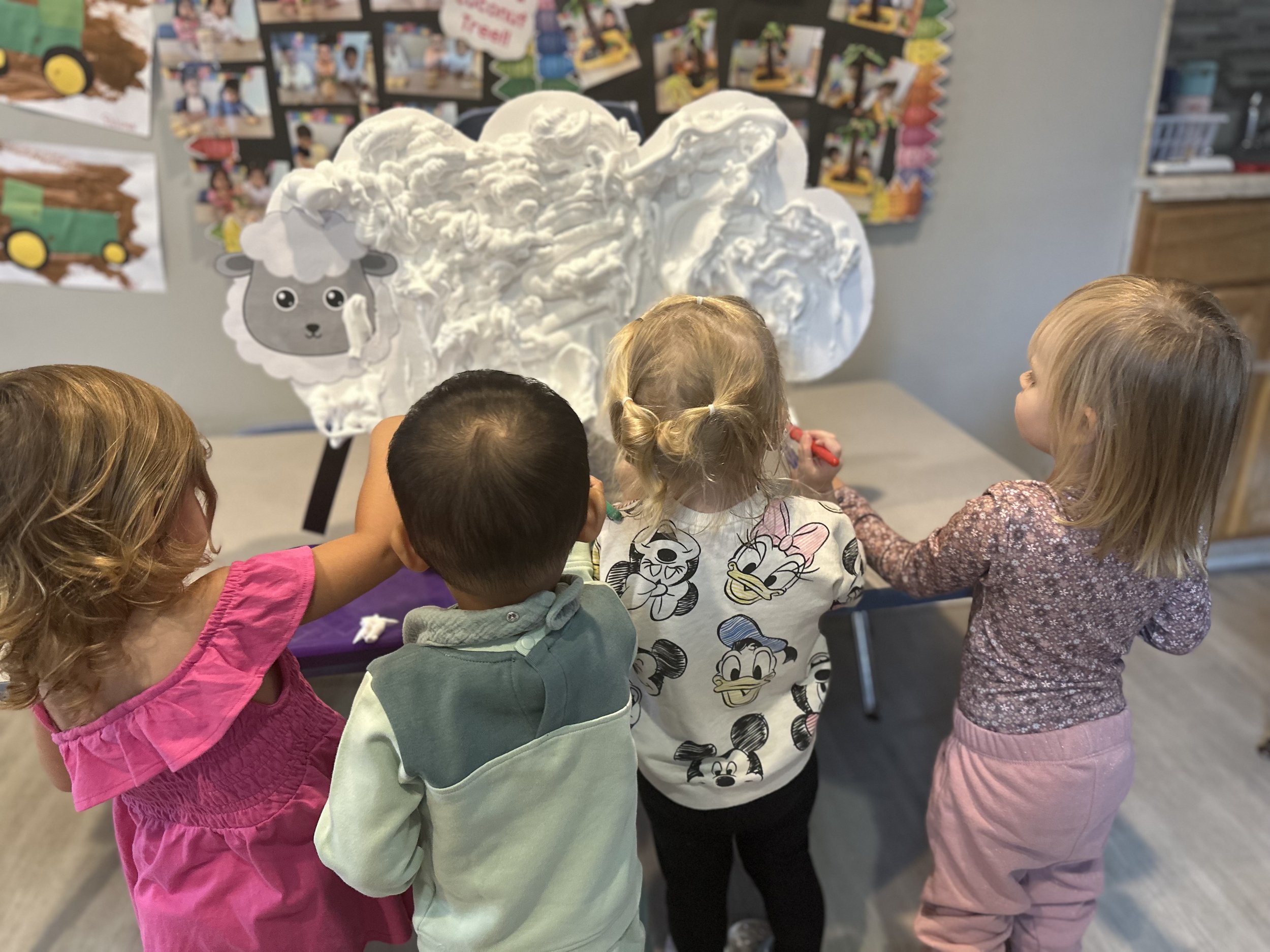 Four children stand around a large, white cotton cloud display with a gray cartoon sheep's face on it, drawing or writing on it in a classroom.