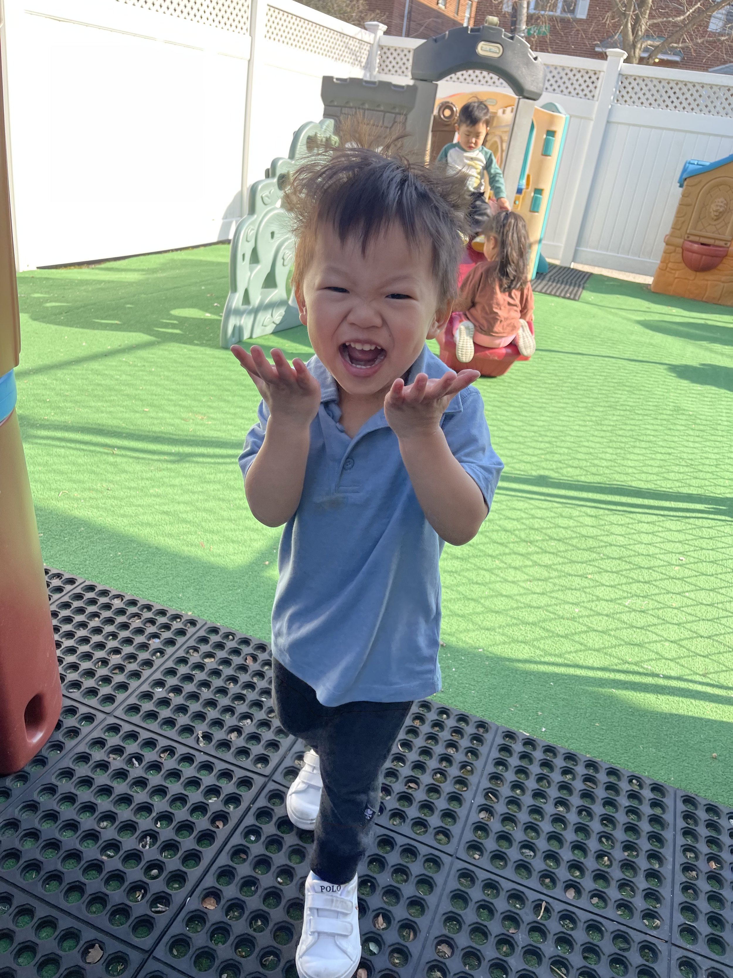 A young boy with messy hair smiling and laughing at a playground, with other children in the background playing on a slide and a swing.
