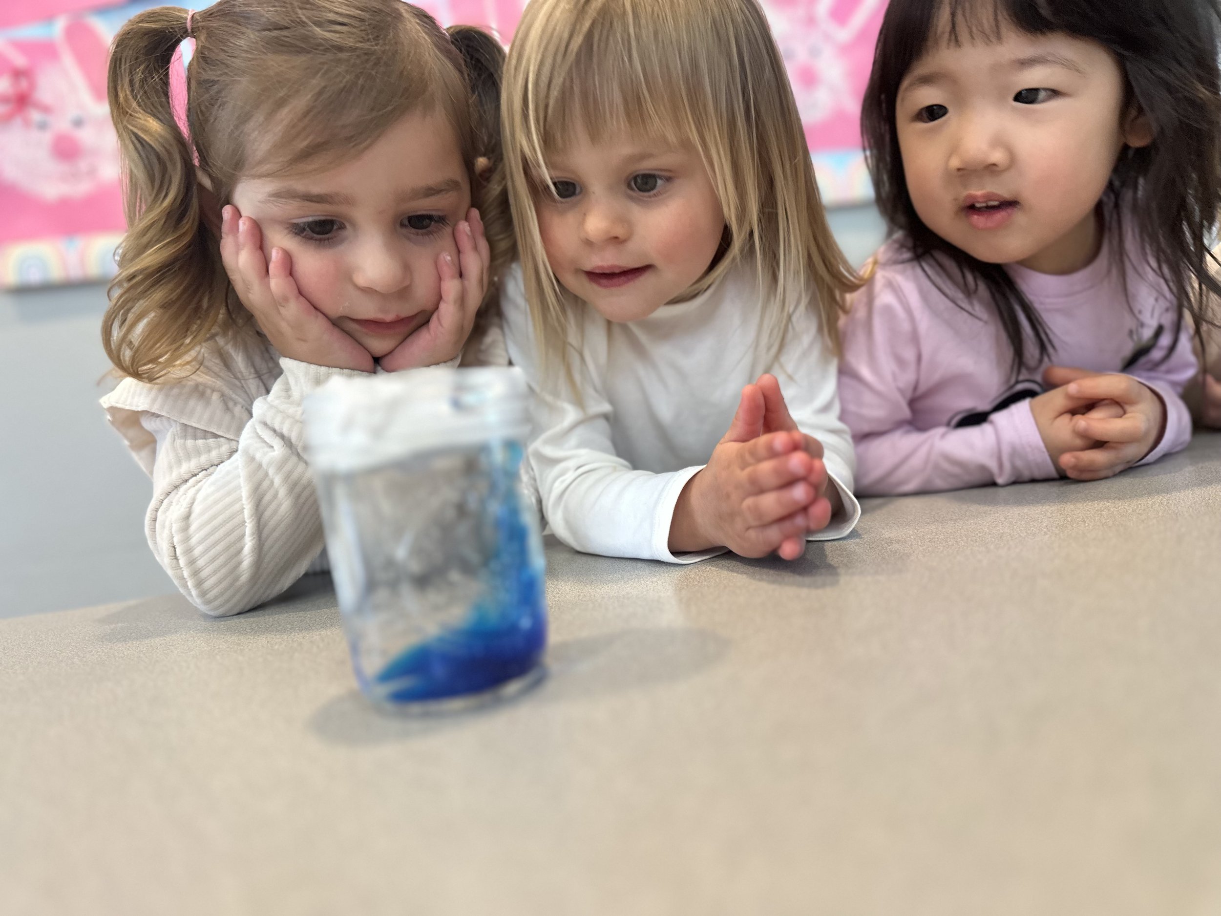 Three young girls observing a small glass jar with blue and white objects on a tabletop.