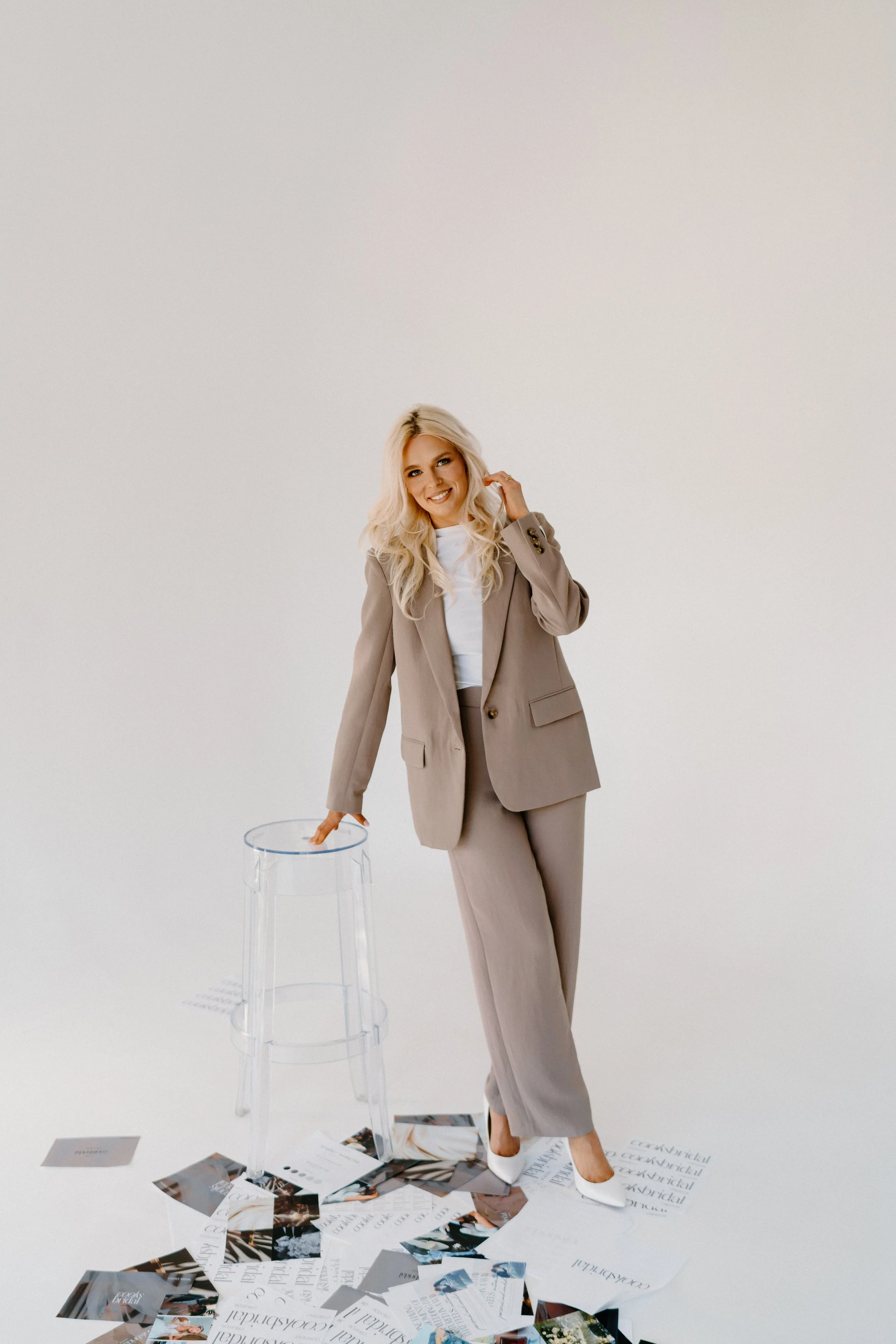 A woman with blonde hair in a light-colored pantsuit standing next to a transparent stool with scattered magazines and papers on the floor, smiling at the camera against a plain white background.