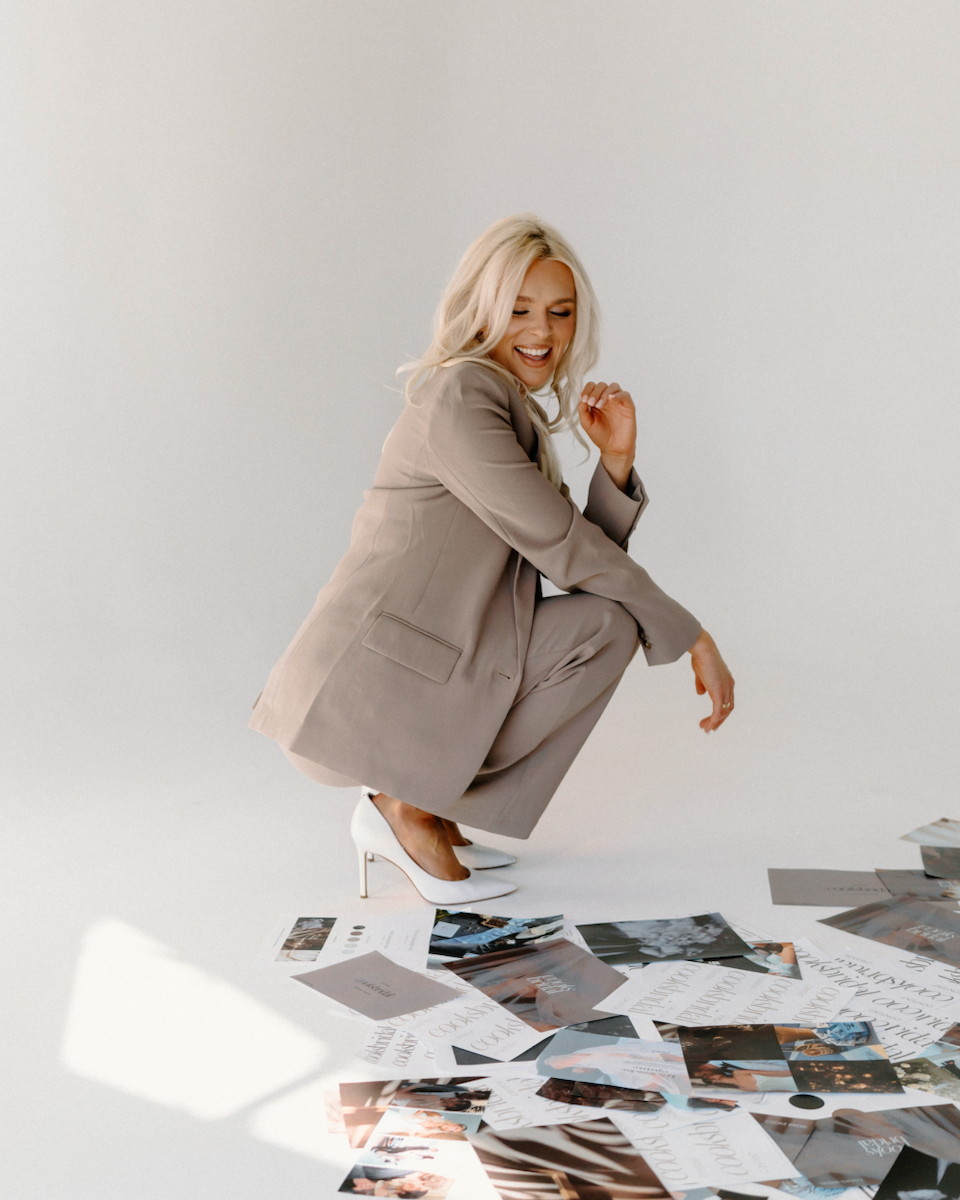 A blonde woman in a beige suit and white high heels is squatting on the floor, smiling and looking down at scattered photographs and papers.