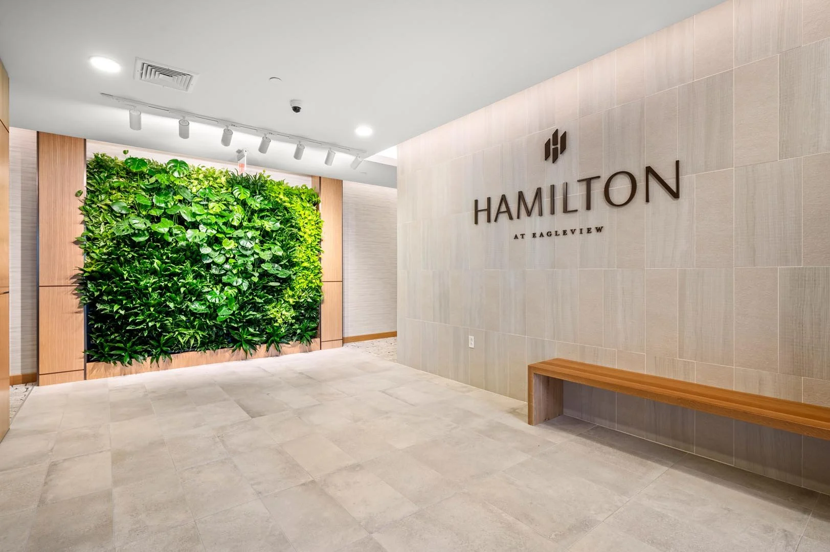 Modern interior space with a vertical green plant wall, tiled walls, overhead lighting, a wooden bench, and "Hamilton at Eagleview" signage.