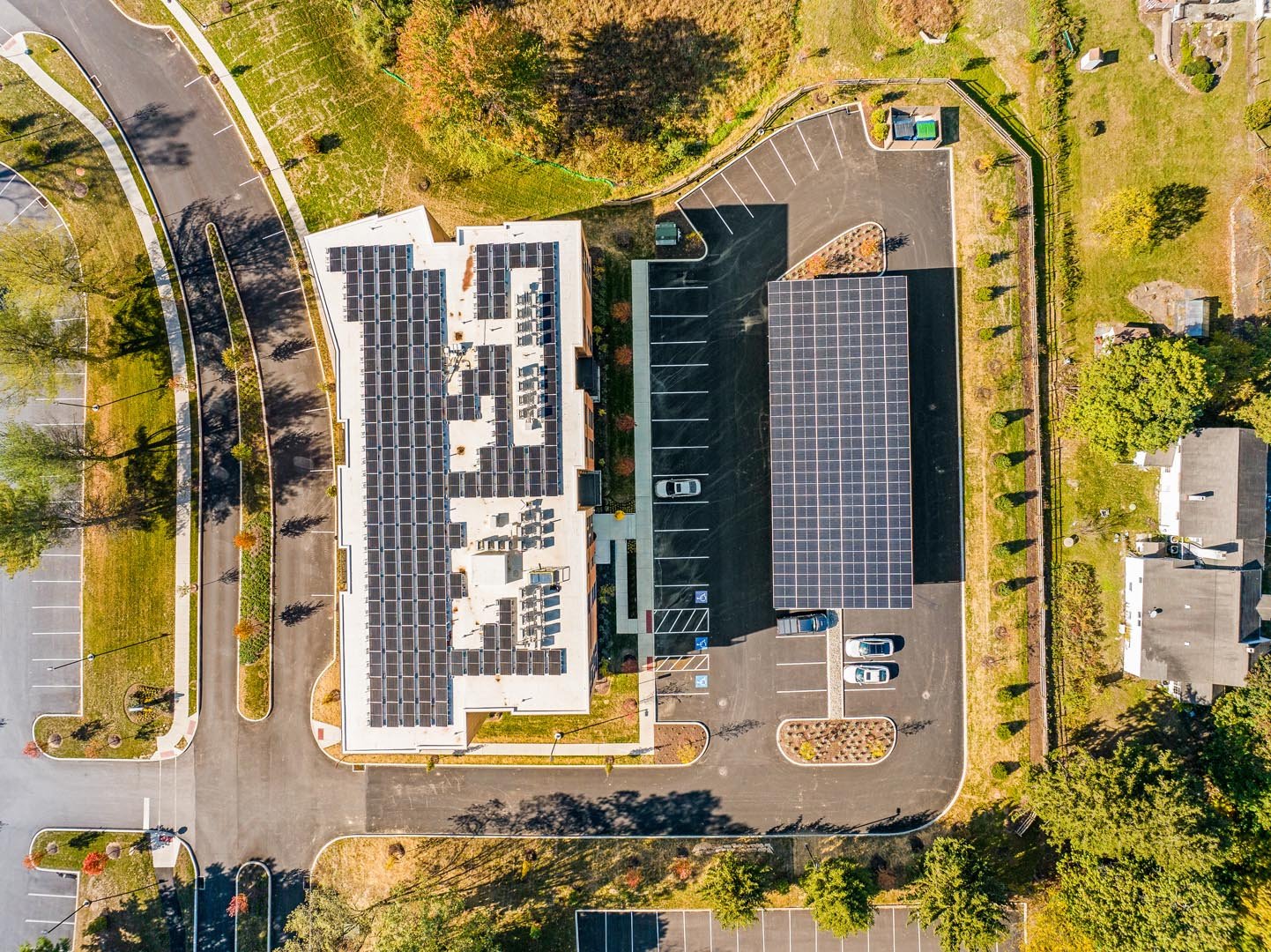 Aerial view of a building with solar panels on the roof and a parking lot, surrounded by greenery.