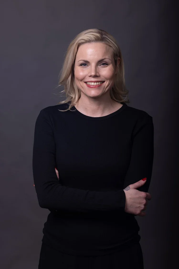 Hän on nuori nainen, jolla on vaalea, suora hiustyyli ja hän hymyilee leveästi. Hänellä on päällä musta pitkähihaisella t-paita ja hän seisoo harmaalla taustalla.