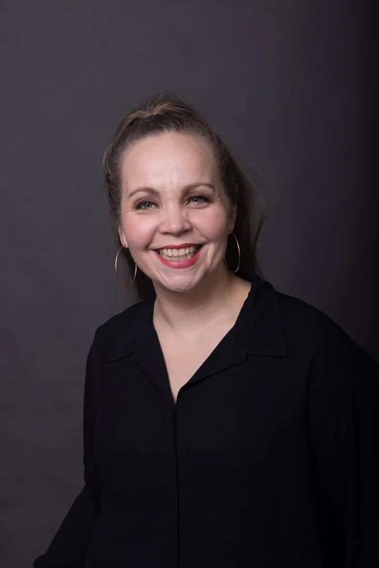 Hymyilevä nainen, jolla on suuri hopeanhohtoinen rengas korvissään, musta pusero ja ruskea kampaus hännässä, seisoo tummanharmaalla taustalla.