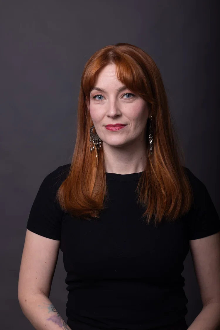 Kuva naisesta, jolla on punaiset hiukset, musta paita ja roikkuvat korvakorut, kohtalaisella taustalla.