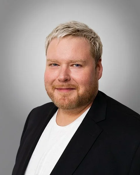 Henkilön muotokuva valkoista taustaa vasten, hänellä vaaleat hiukset, pienen parta ja hymy.