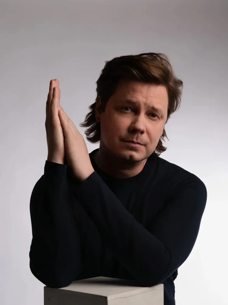 Mies katsoo suoraan kameraan, pitelee kämmentä kasvojensa vieressä, taustalla vaaleanharmaa.