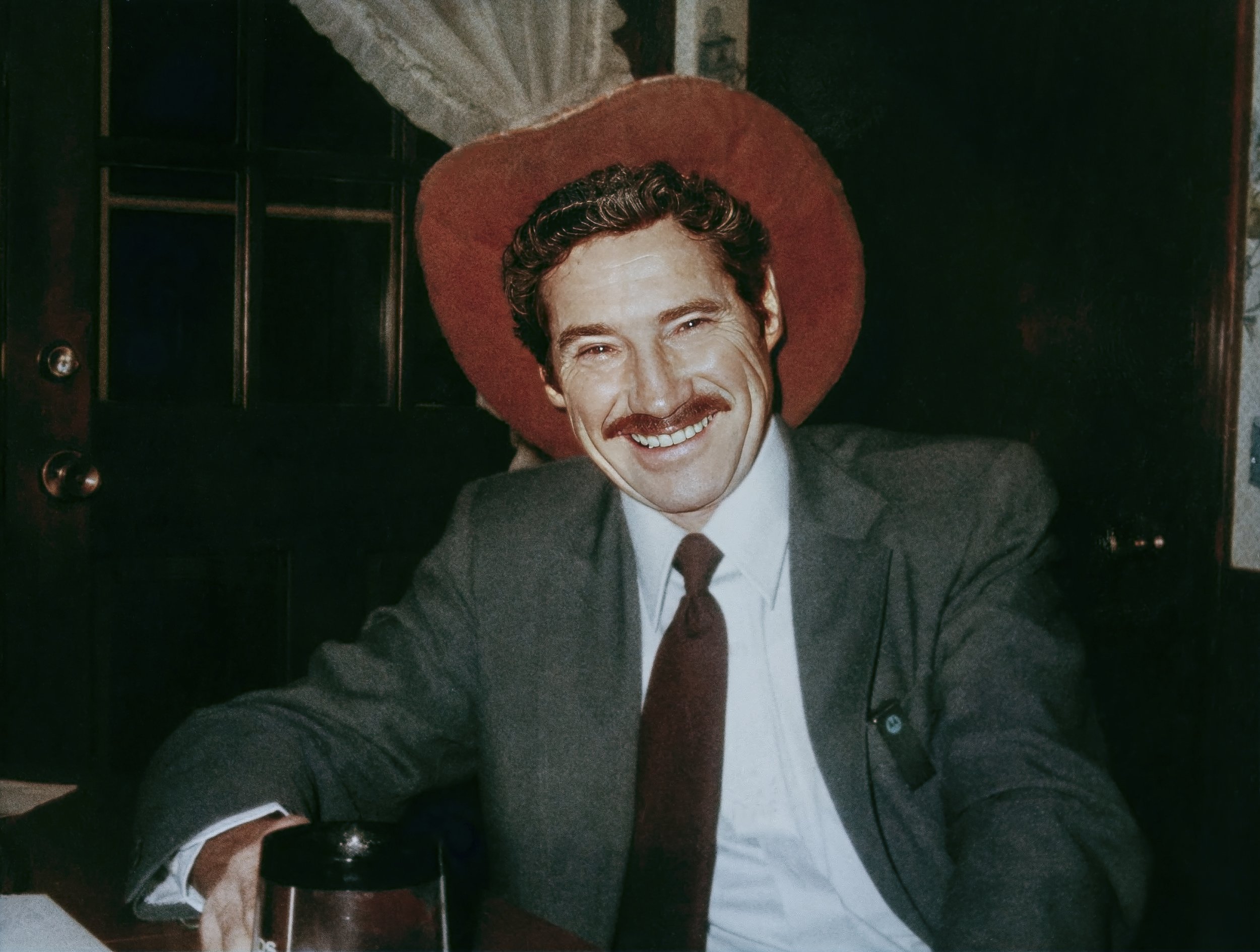 A man wearing a gray suit, white shirt, and red tie, smiling, sitting at a table with a large red hat or cushion behind him.