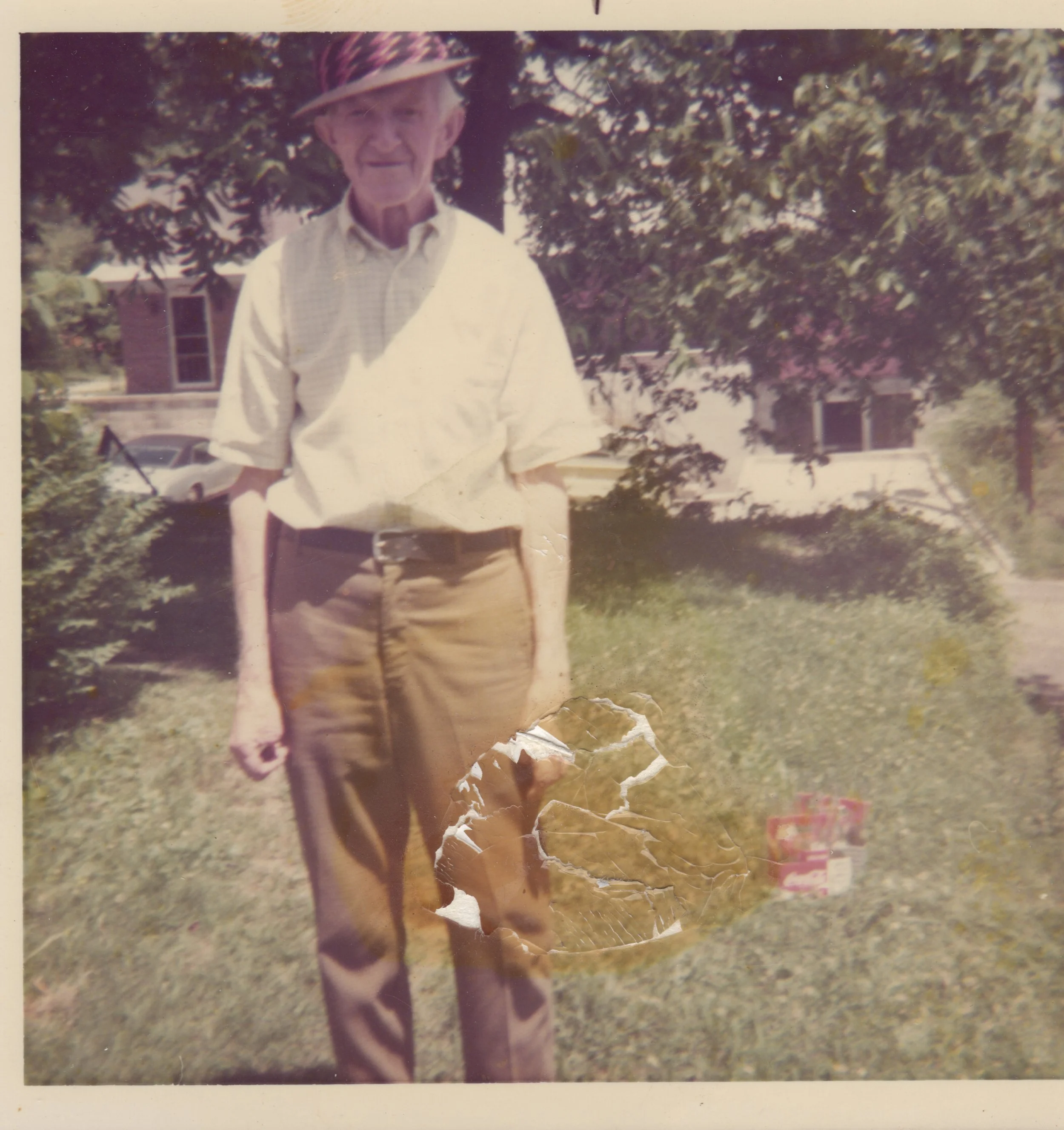An elderly man standing outdoors on grass with trees and buildings in the background. He is wearing a wide-brimmed hat, a light-colored short-sleeved shirt, and brown pants. There is a damaged paper or photo piece near his waist and some boxes on the ground.