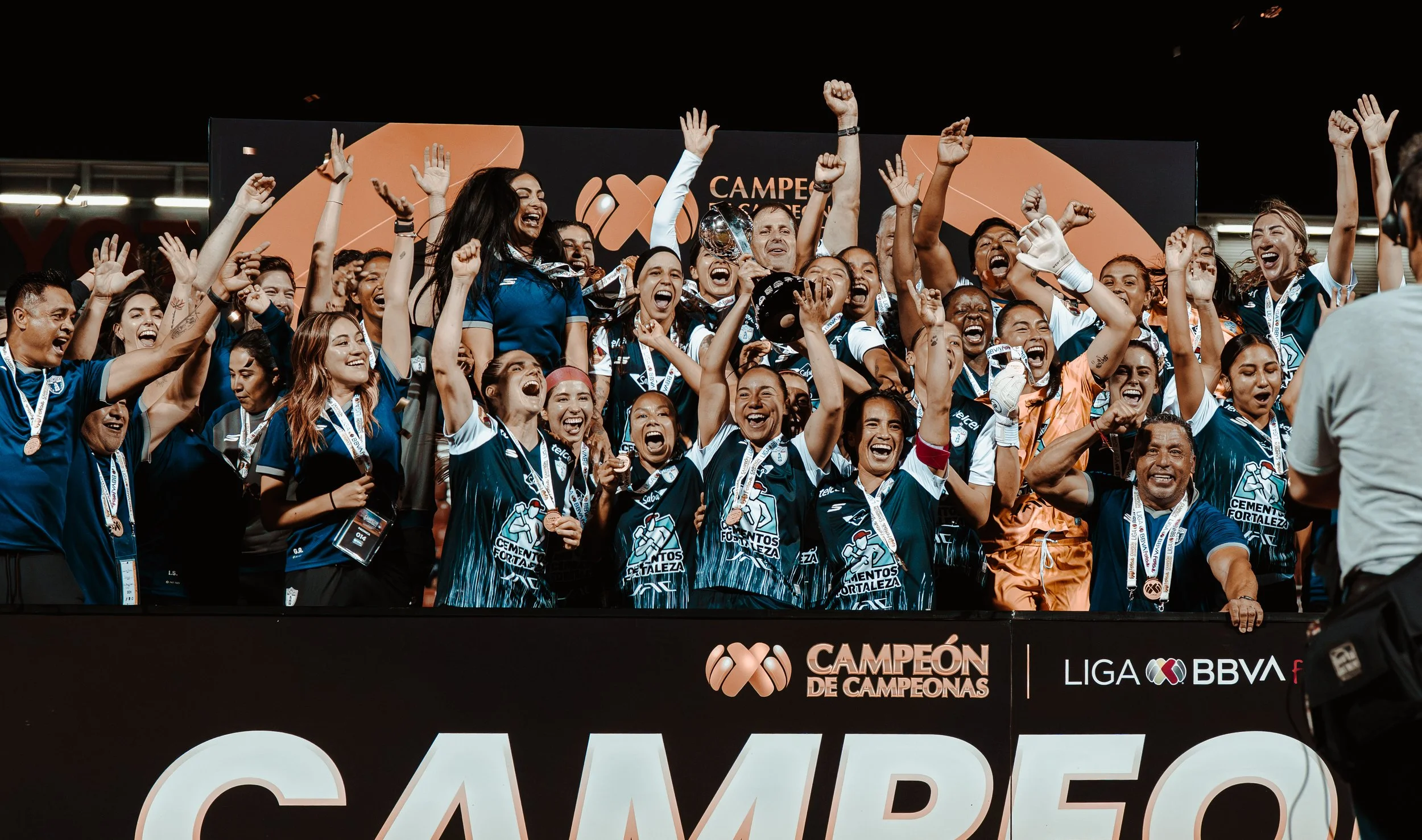 Female soccer Pachuca Femenil team celebrating with medals and trophy after winning championship, on stage with 'Campeón de Campeonas' banner.