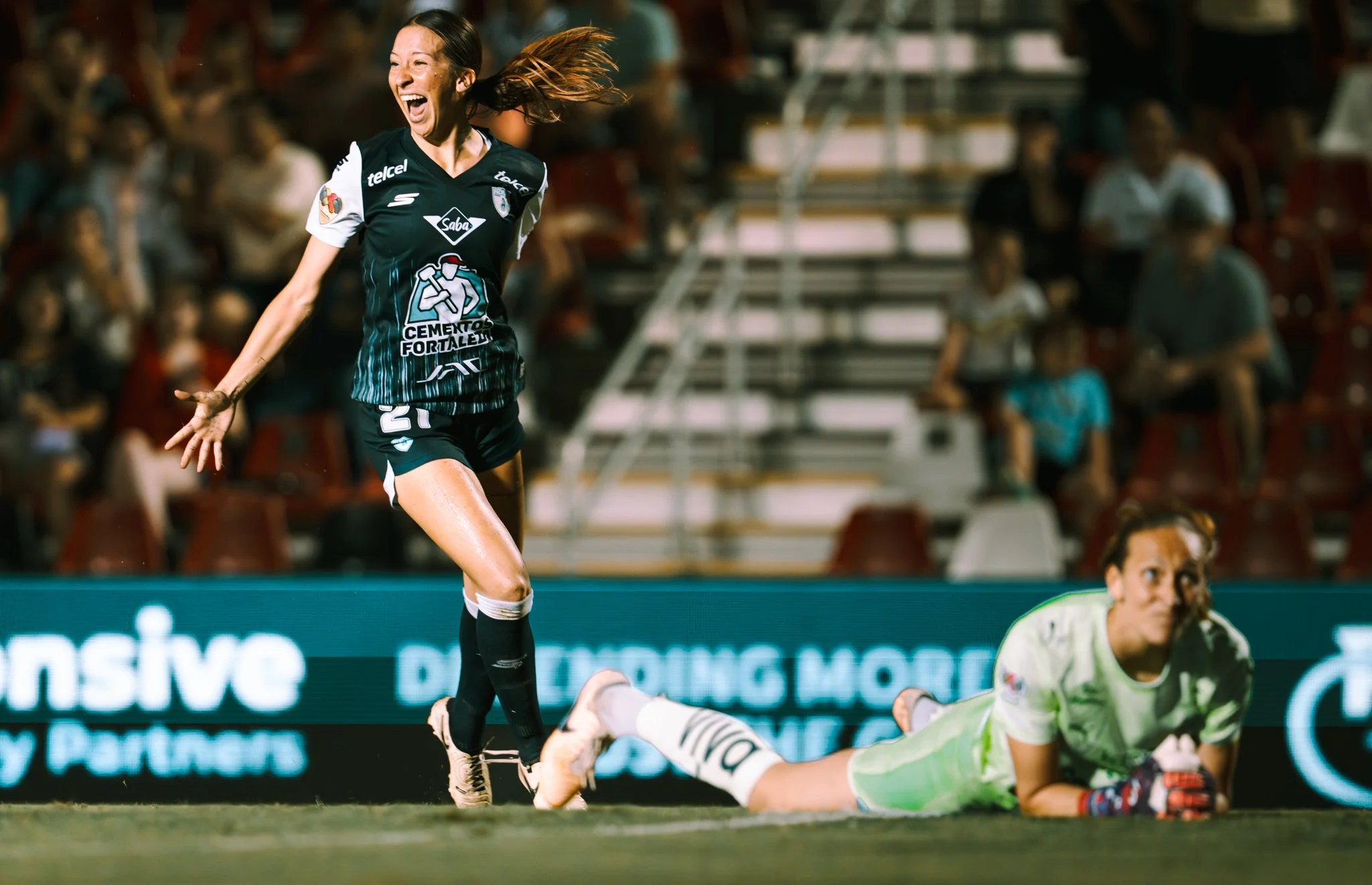 A female soccer player in a black and teal uniform celebrating on the field while a goalkeeper in white and green uniform lies on the ground.