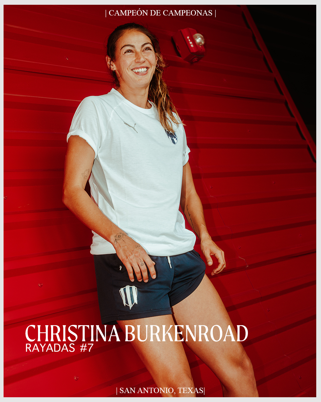 A female soccer player standing against a red wall, smiling, wearing a white sports shirt and black shorts with team logos, in San Antonio, Texas.
