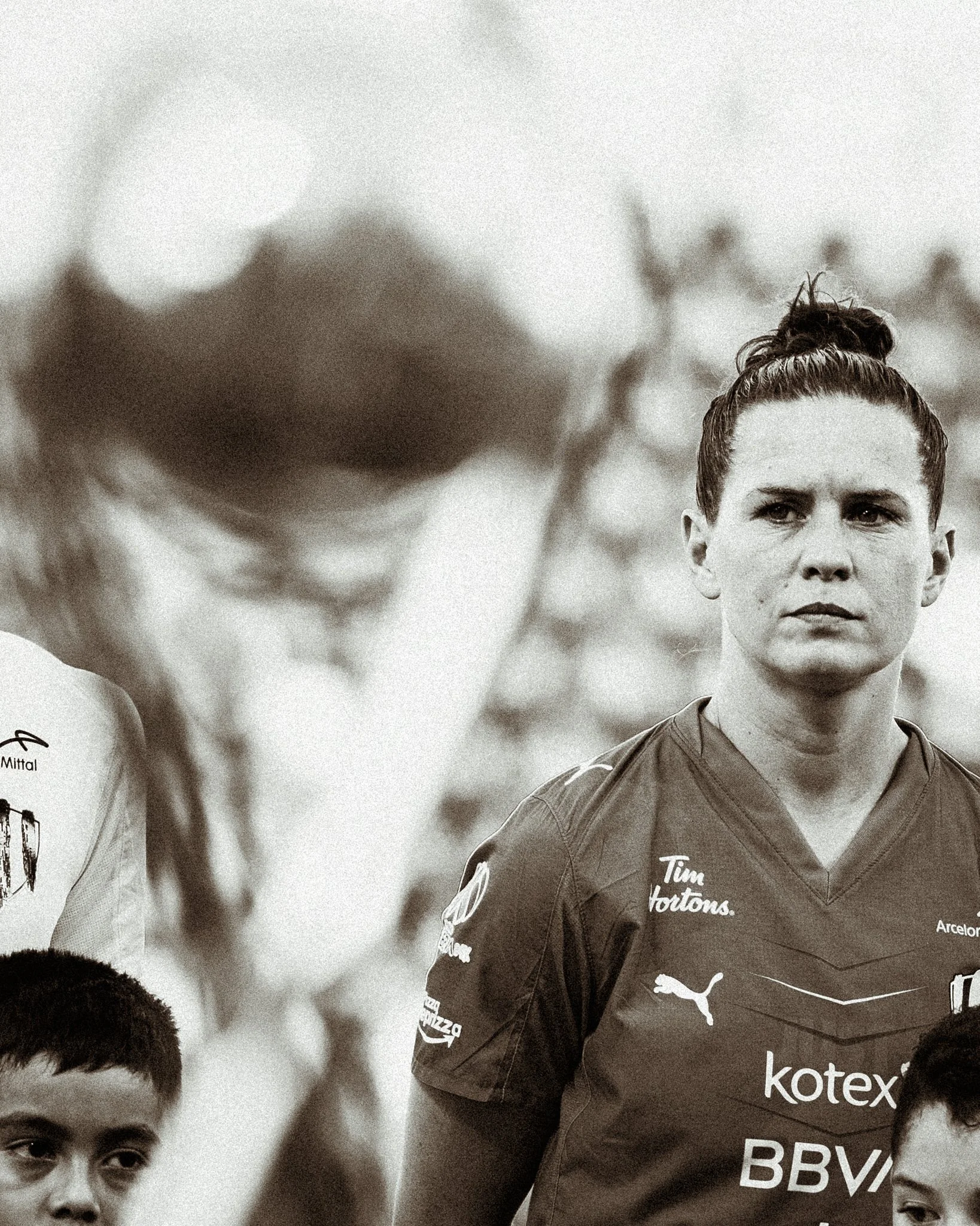A woman with a bun hairstyle wearing a sports jersey standing outdoors with children nearby.
