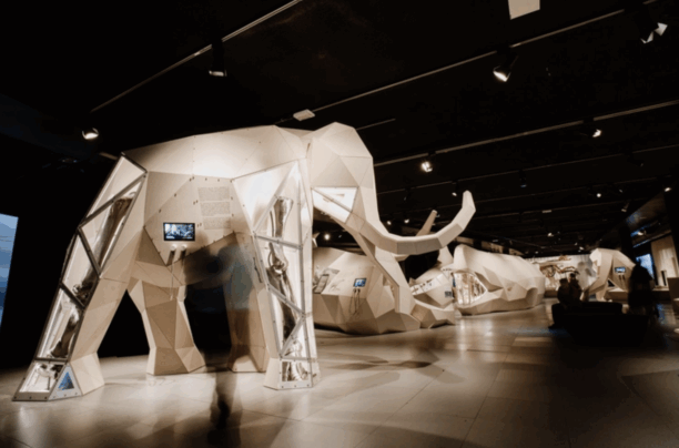 Indoor exhibit of geometric, white, animal-shaped structures resembling oxen or buffalo, with some display screens, in a dark room with visitors.