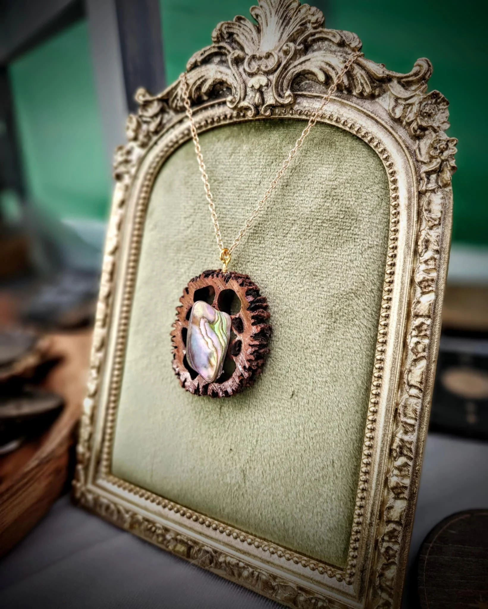A decorative necklace with a large, iridescent crystal pendant on a walnut slice on a gold chain, displayed on a vintage ornate gold frame with green fabric background.