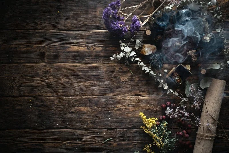 Arrangement of dried flowers, herbs, and crystals on a rustic wooden surface with some glass bottles and smoke.