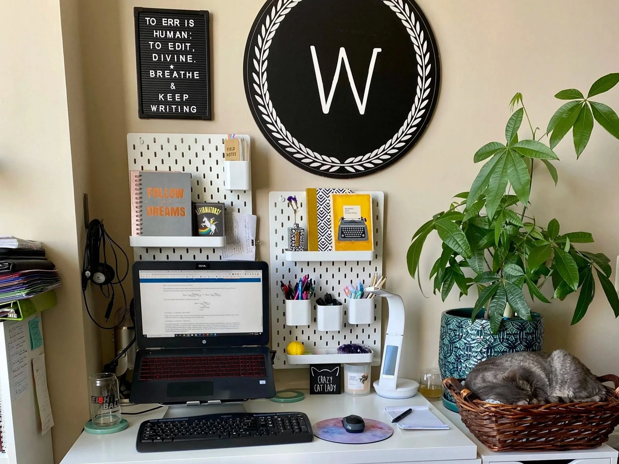 A writer's desk setup with a letter board reading "To err is human; to edit, divine. Breathe and keep writing," a W monogram sign, pegboard with notebooks and writing supplies, a laptop open to a document, and sleeping cat in a basket beside a plant.
