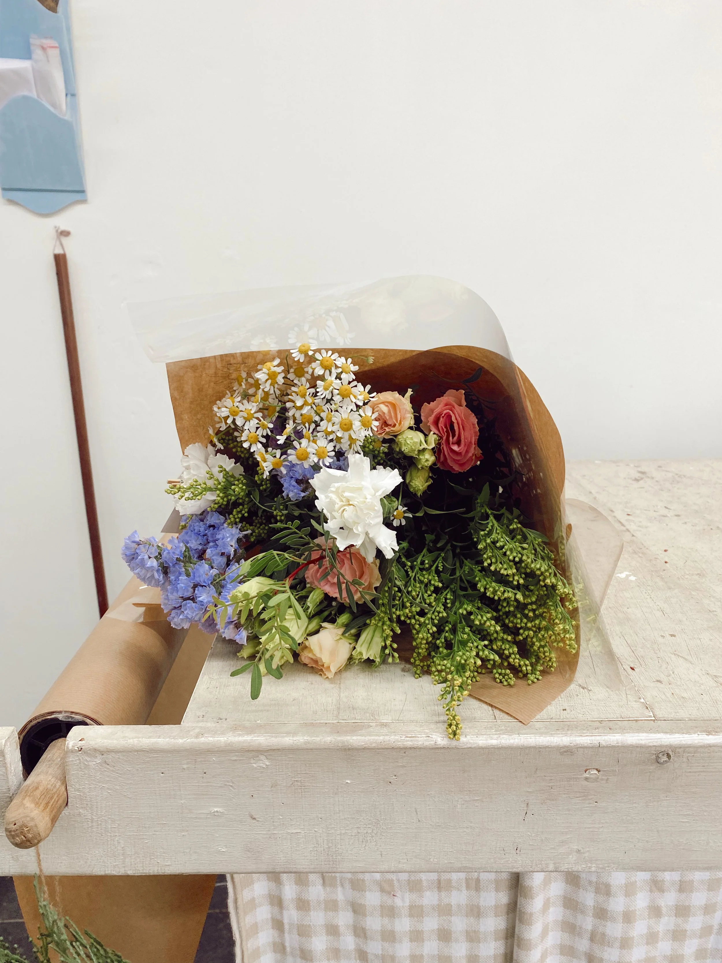 A bouquet of various colorful flowers, including daisies, roses, and other blooms, wrapped in brown paper and transparent plastic, resting on a white wooden table.