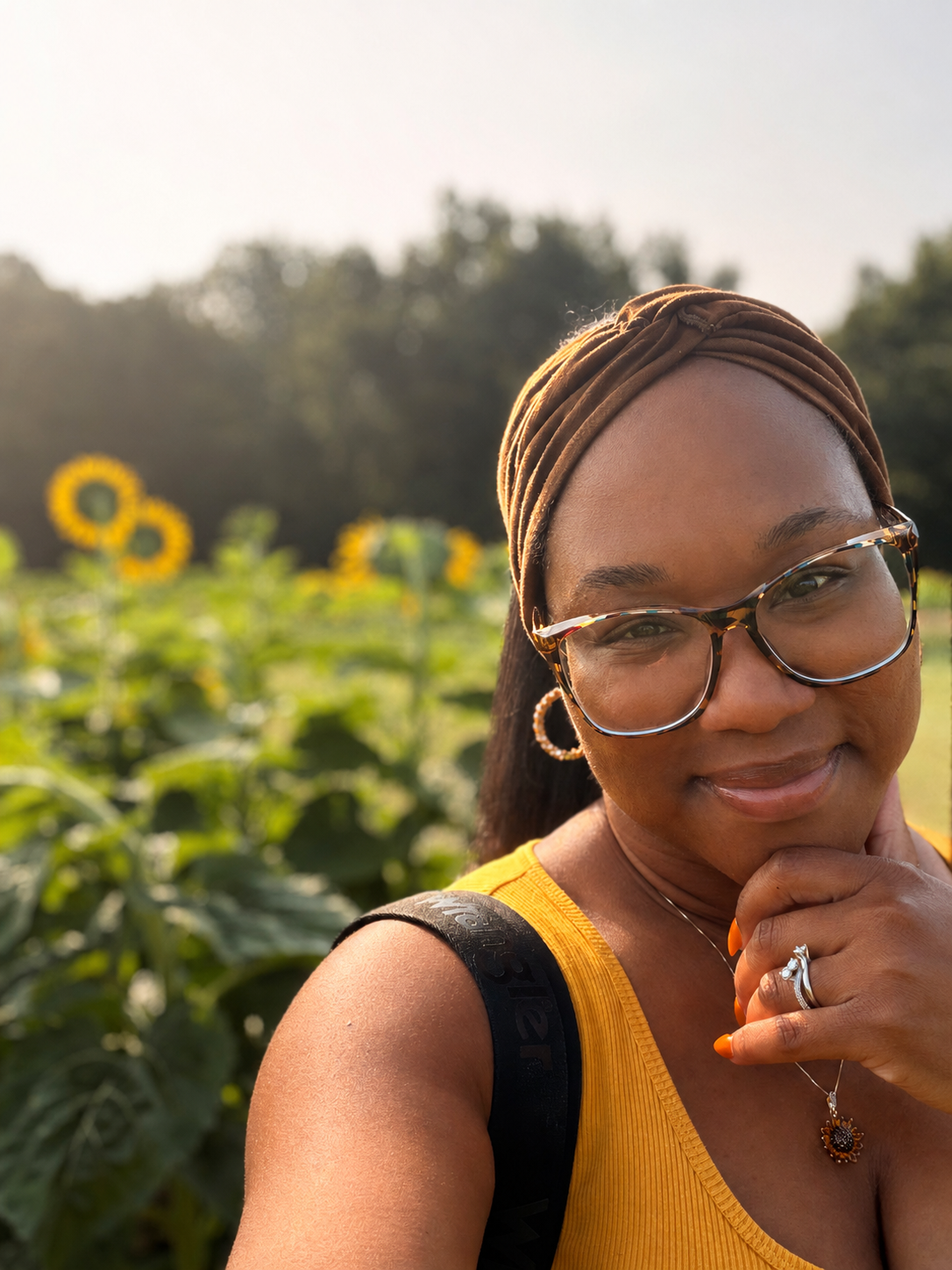 A woman with glasses and earrings takes a selfie outdoors amidst a sunflower field, smiling gently.