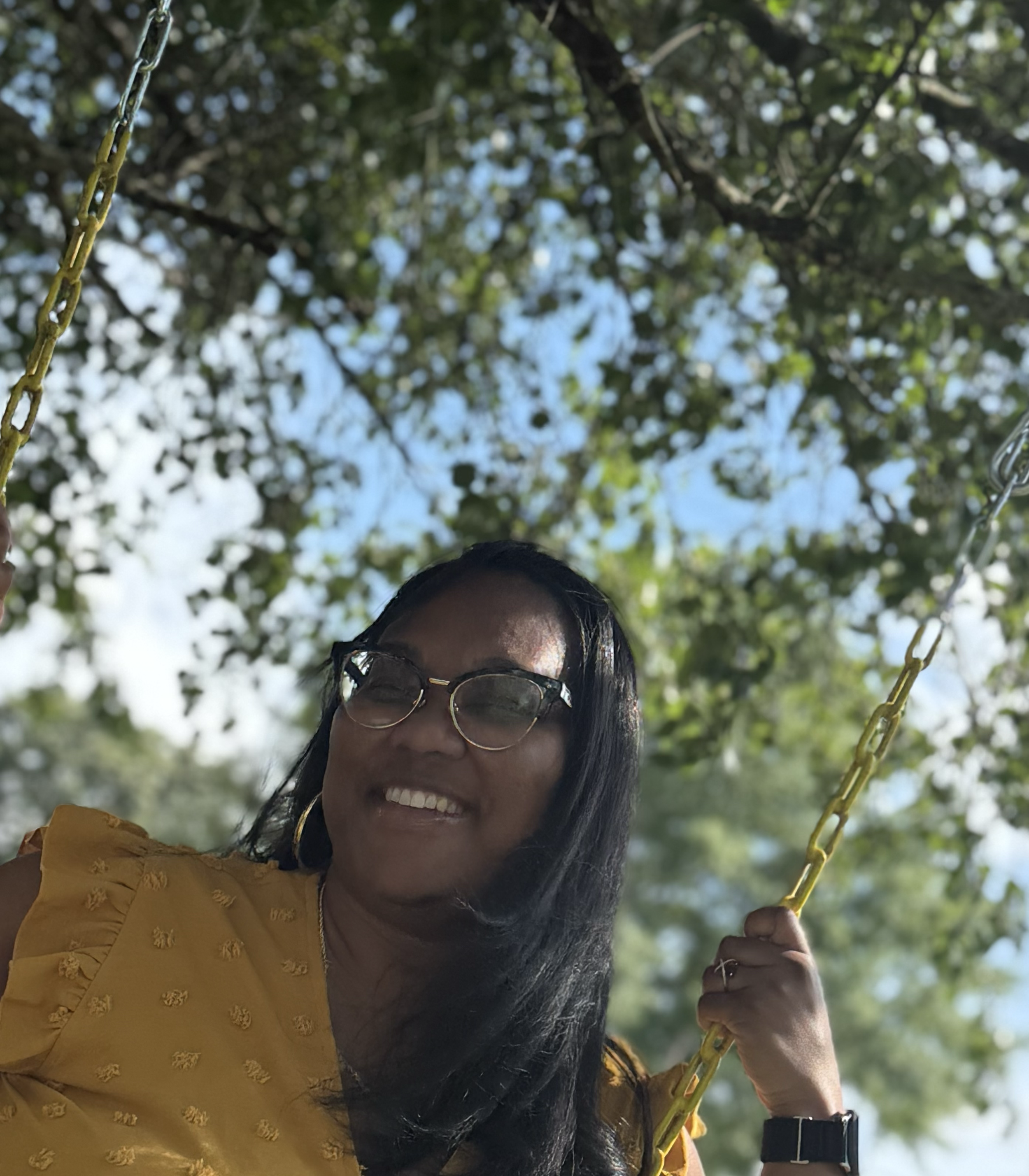 A woman smiling on a swing, wearing sunglasses and a yellow top, with a background of trees and blue sky.