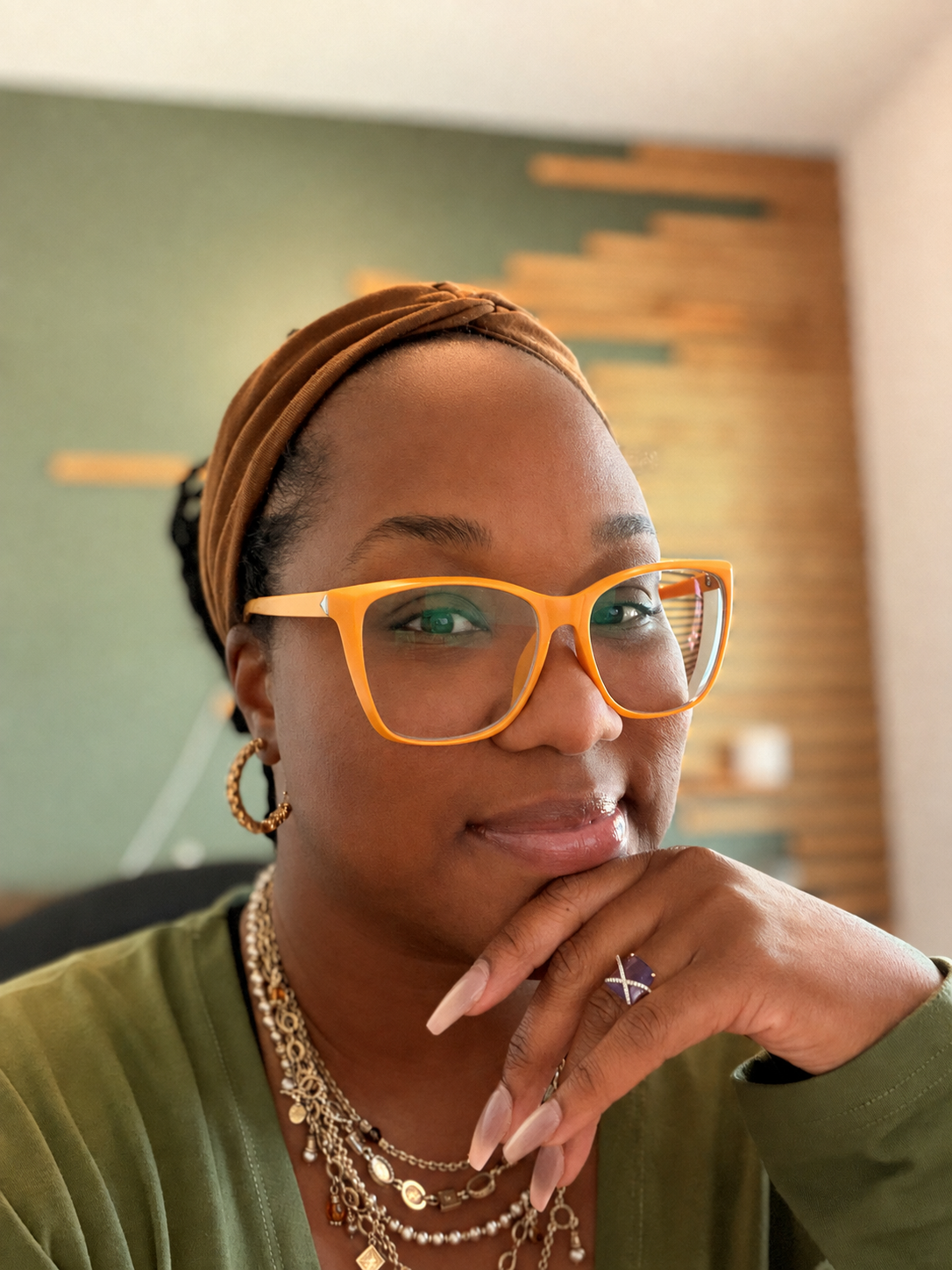 A woman wearing orange glasses, gold hoop earrings, layered necklaces, and a brown headband, resting her chin on her hand and smiling softly.