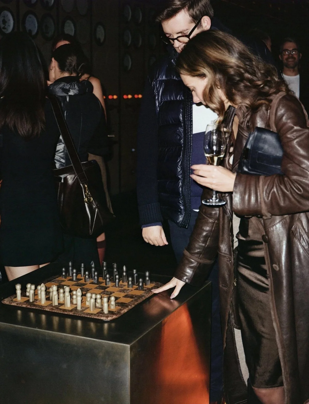 Two people playing chess at a table, one woman is moving a chess piece, and another holding a glass of wine in her hand.