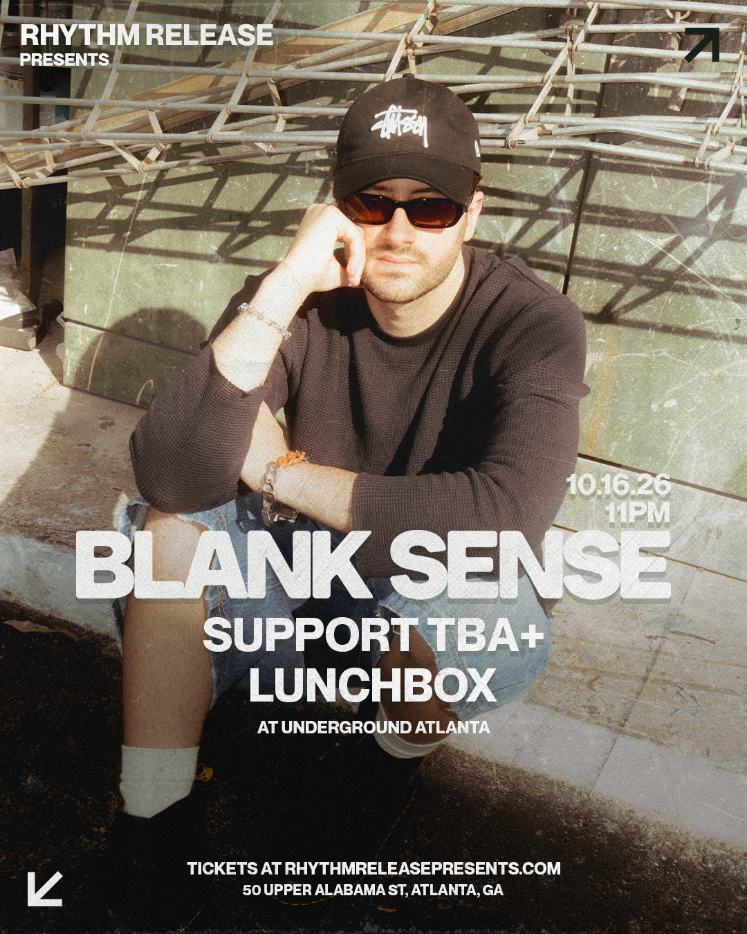 A young man wears sunglasses, a black cap and black shirt, sitting outside on the sidewalk with a serious expression, with event details overlaid on the image for a music event.