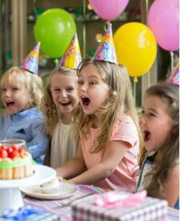 Four young girls celebrating a birthday party with colorful balloons and wearing party hats, sitting at a decorated table with a cake.