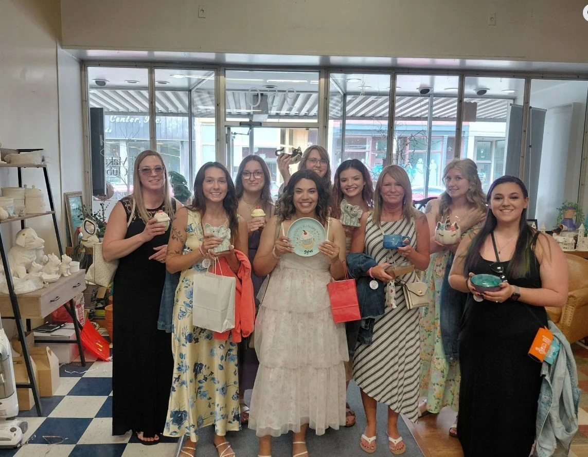 A group of ten women attending a bridal shower, smiling and holding dishes and cups inside a shop that sells pottery and gift items on a walk-in basis.