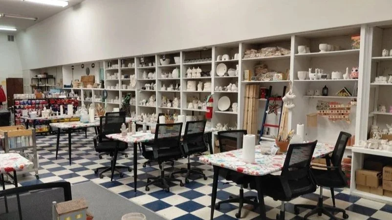Room with shelves filled with unpainted ceramic figurines and dishes, tables covered with colorful tablecloths, and chairs, set up for a ceramics painting activity or paint-your-own Pottery Class.